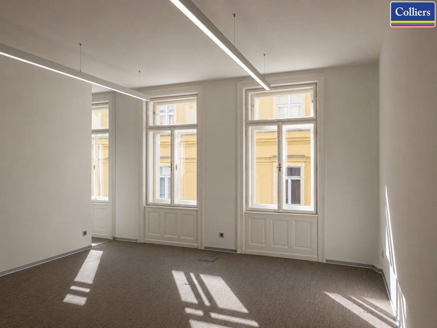 Geräumiger Büroraum mit Teppichboden und hohen Fenstern, die für eine helle und offene Arbeitsumgebung sorgen.
