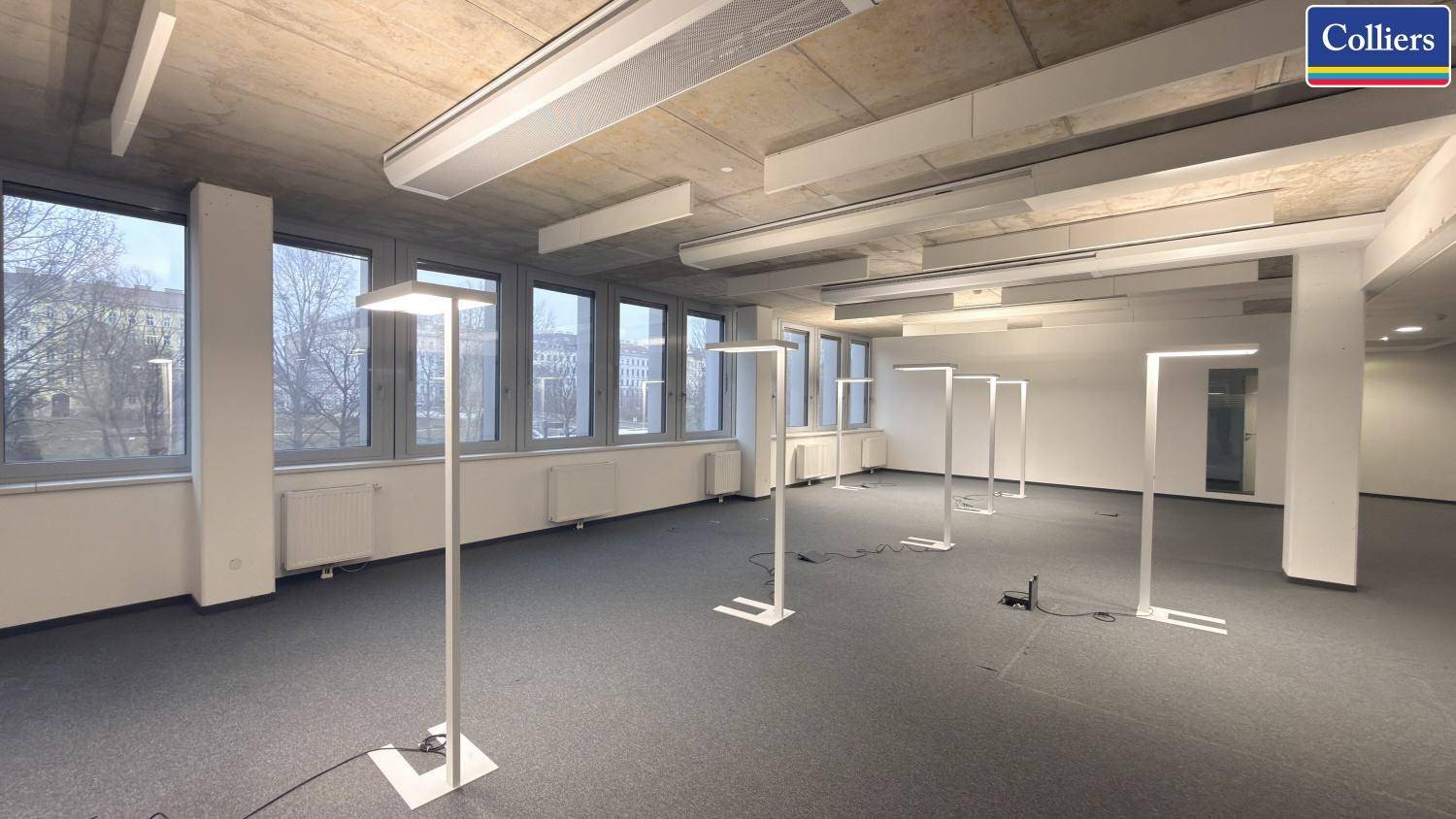 Geräumiges Büro mit Teppichboden, durchgehenden Fenstern und modernen Stehleuchten, die den Raum erhellen.