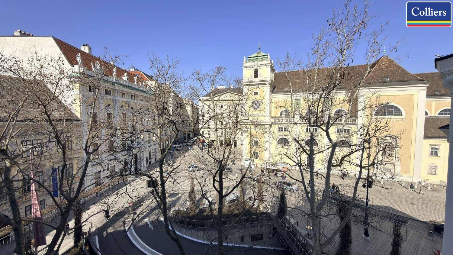 Historische Stadtansicht mit einem großen Platz, umgeben von Gebäuden und einem Kirchturm unter blauem Himmel.