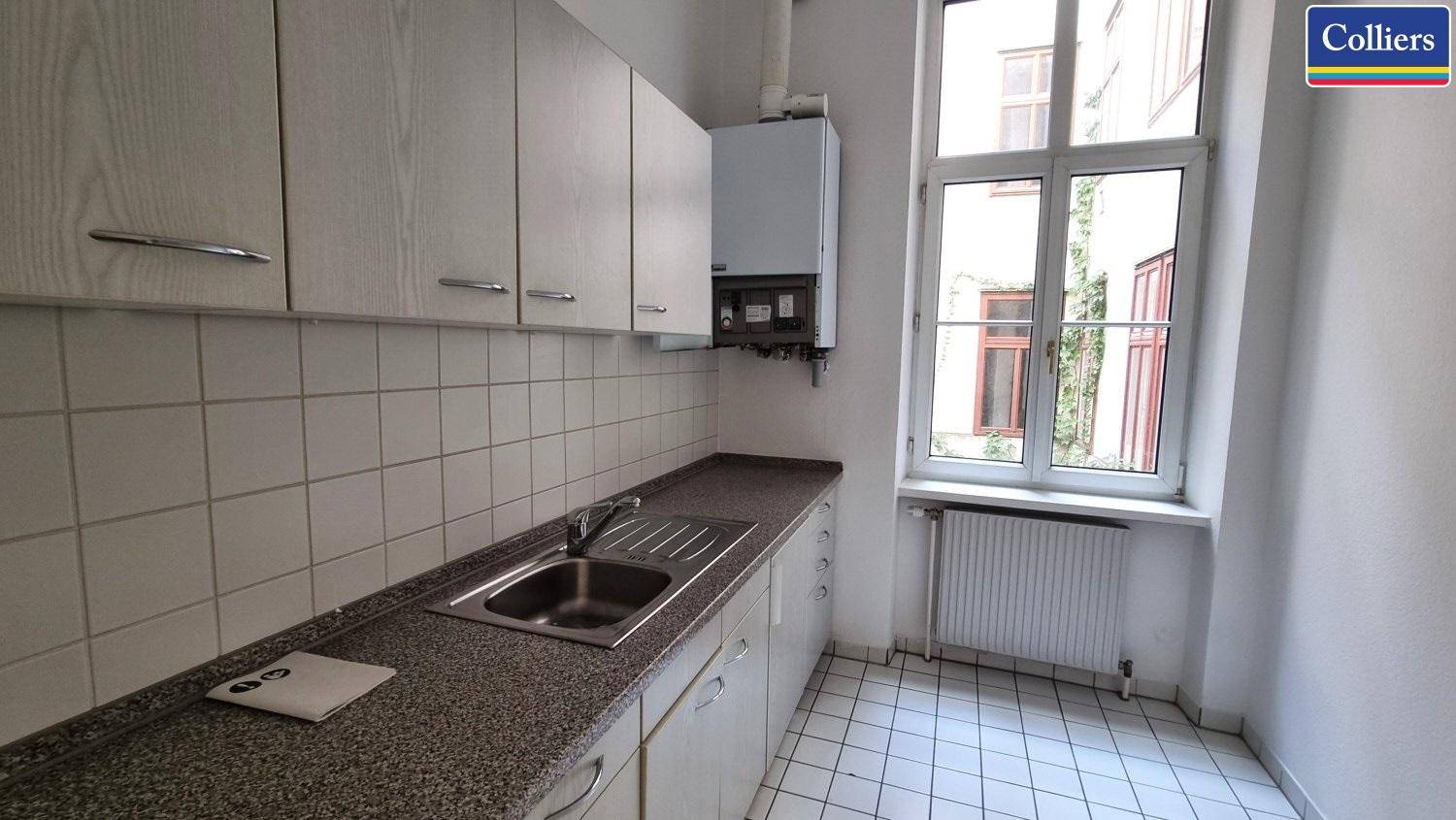 Eine kleine Küche mit weißen Fliesen, hellen Schränken und einem Fenster mit Blick auf einen Innenhof.