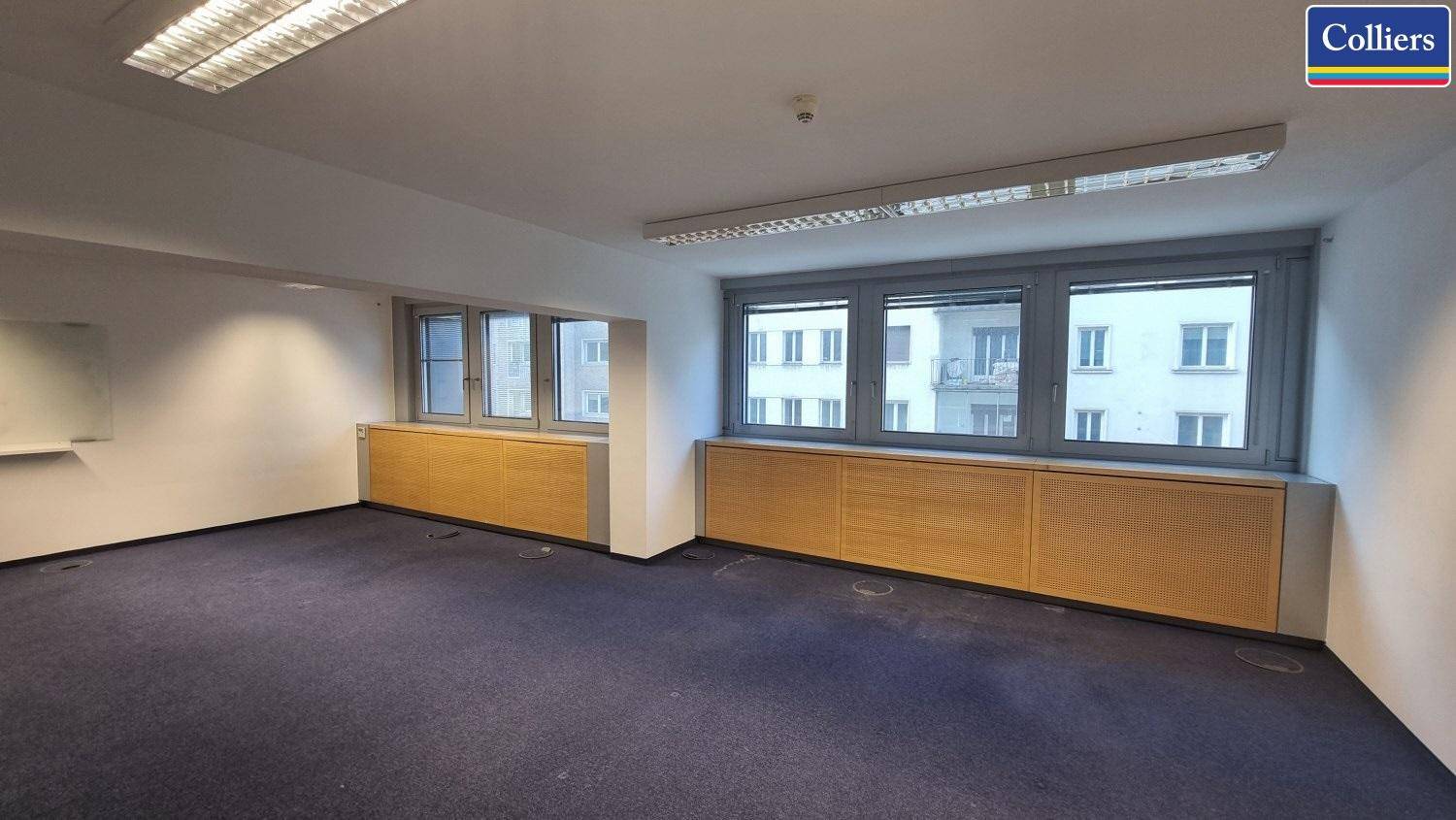 Flexibler Büroraum mit Teppichboden, breiten Fenstern und integrierten Holzschränken.