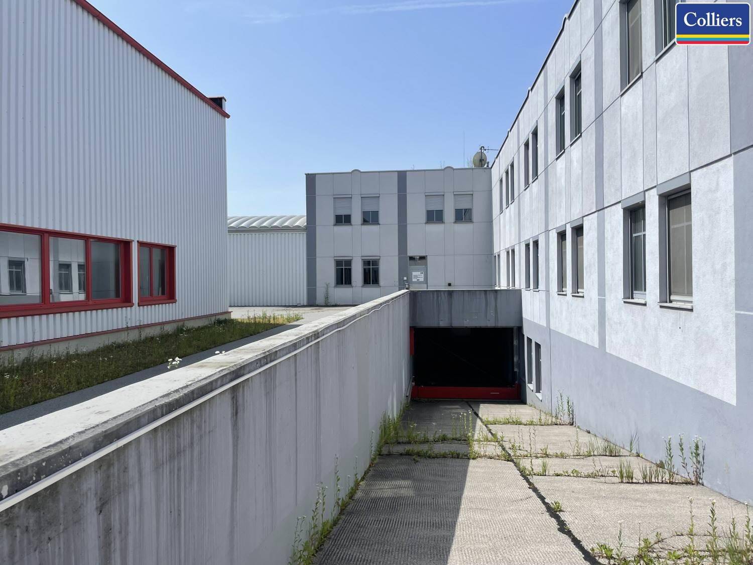 Zufahrt zu einer Tiefgarage zwischen zwei Gebäudeteilen mit Betonwänden und wenig Vegetation.