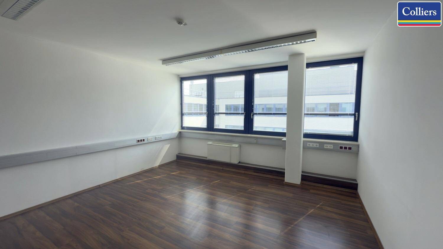 Bürofläche mit Holzboden und einer Fensterreihe, die einen Blick auf ein gegenüberliegendes Gebäude bietet.