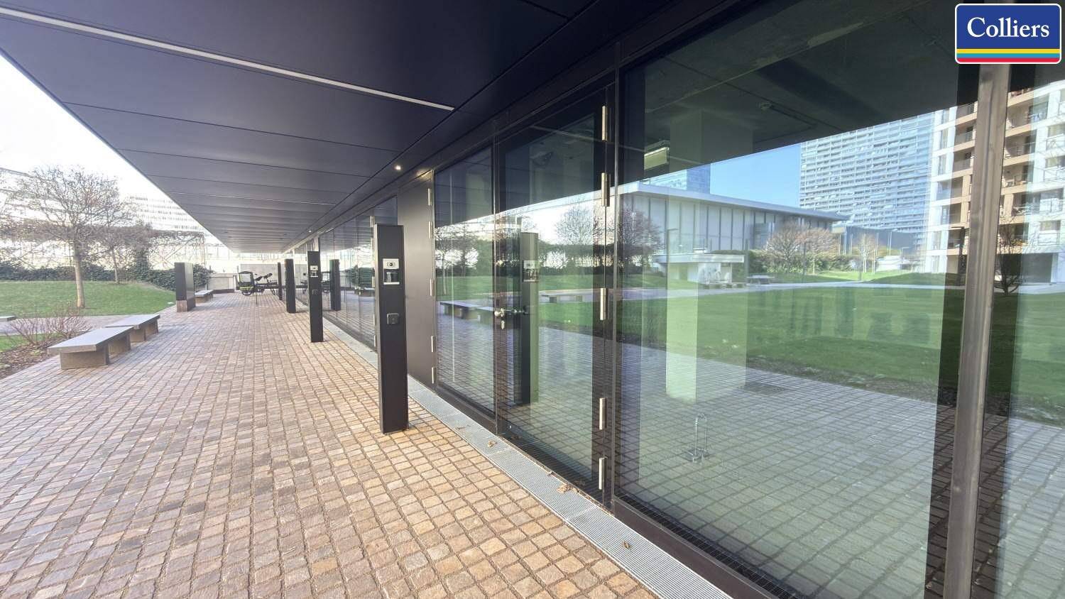 Lange, moderne Glasfassade eines Geschäftsgebäudes mit einem breiten, gepflasterten Gehweg und Sitzgelegenheiten.