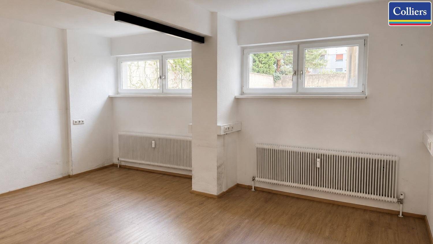 Kleinerer Büroraum mit zwei Fenstern, Heizkörpern und hellem Laminatboden, ideal für Einzelbüros.