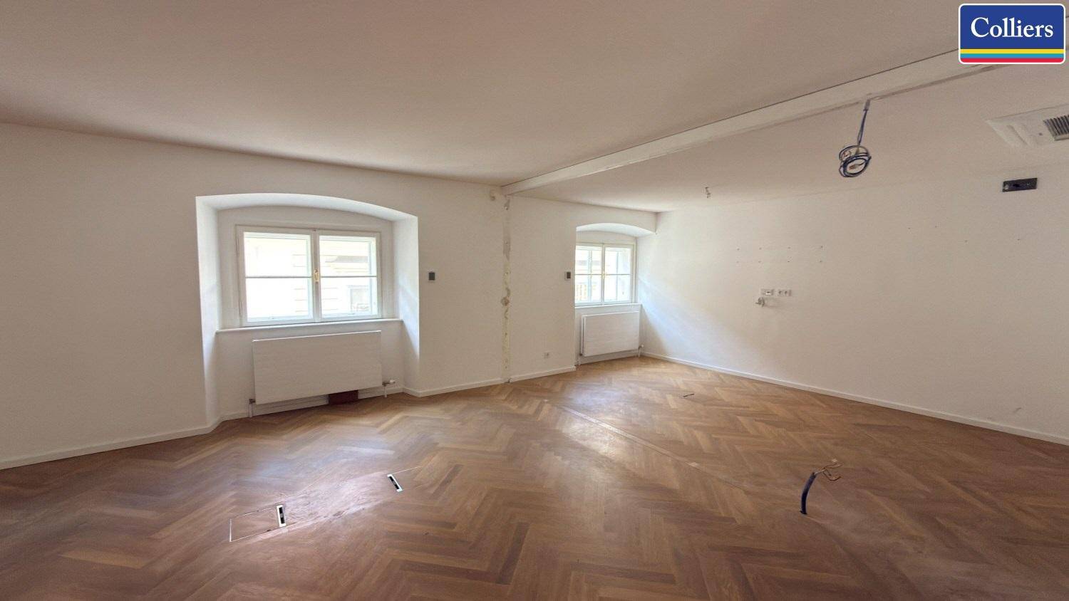 Geräumiger Büroraum mit Fischgrätparkett und Fenstern, die viel Tageslicht hereinlassen.