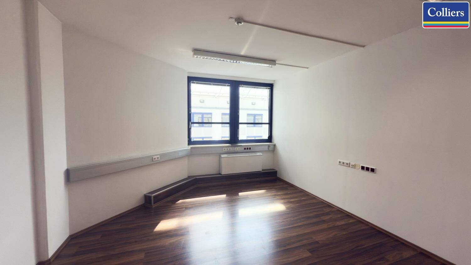 Helles Büro mit Holzboden und einem Fenster, das den Blick auf ein gegenüberliegendes Gebäude freigibt.