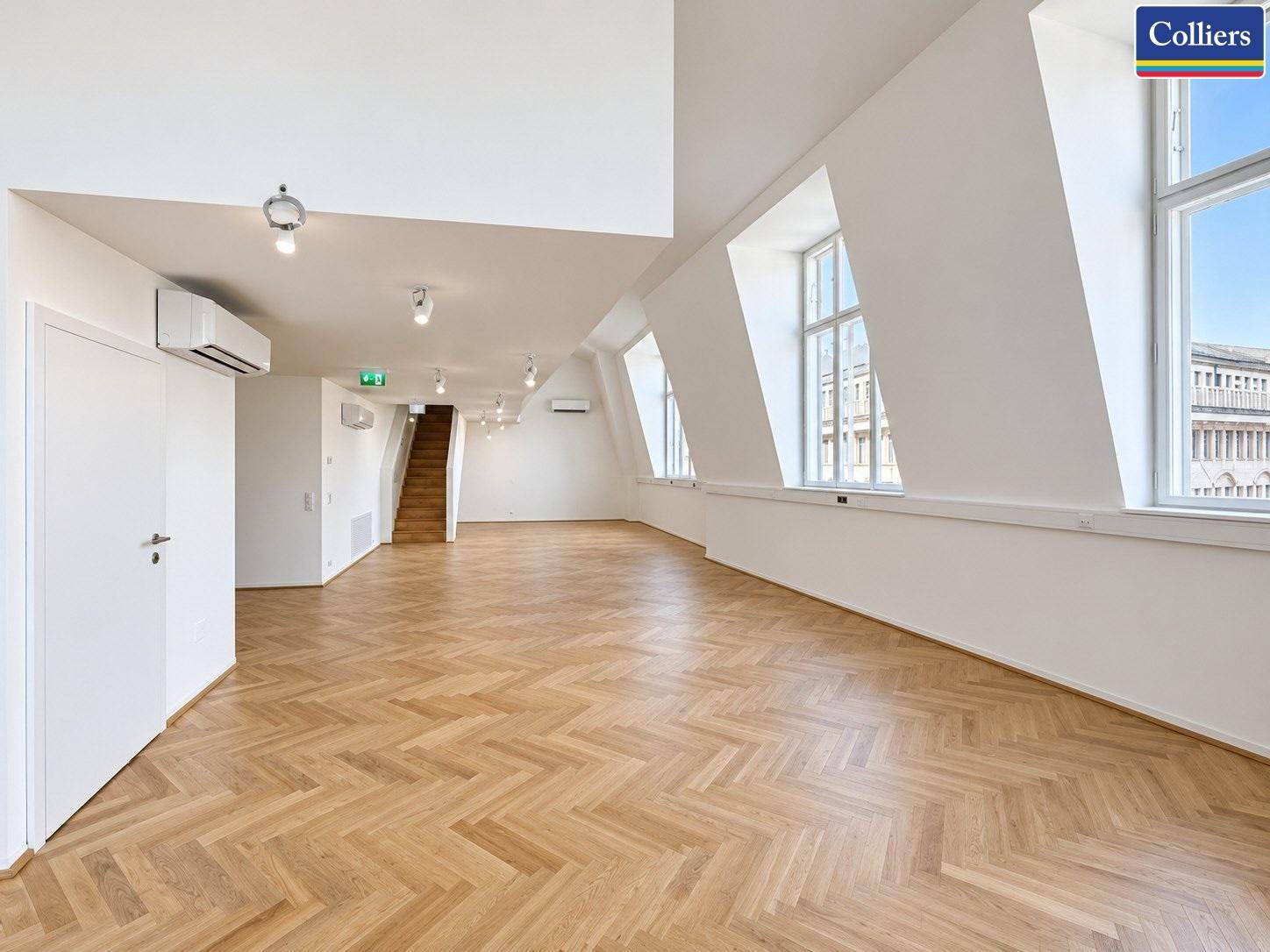 Offener Büroraum mit Fischgrätparkett, Klimaanlage und einer Holztreppe im Hintergrund.