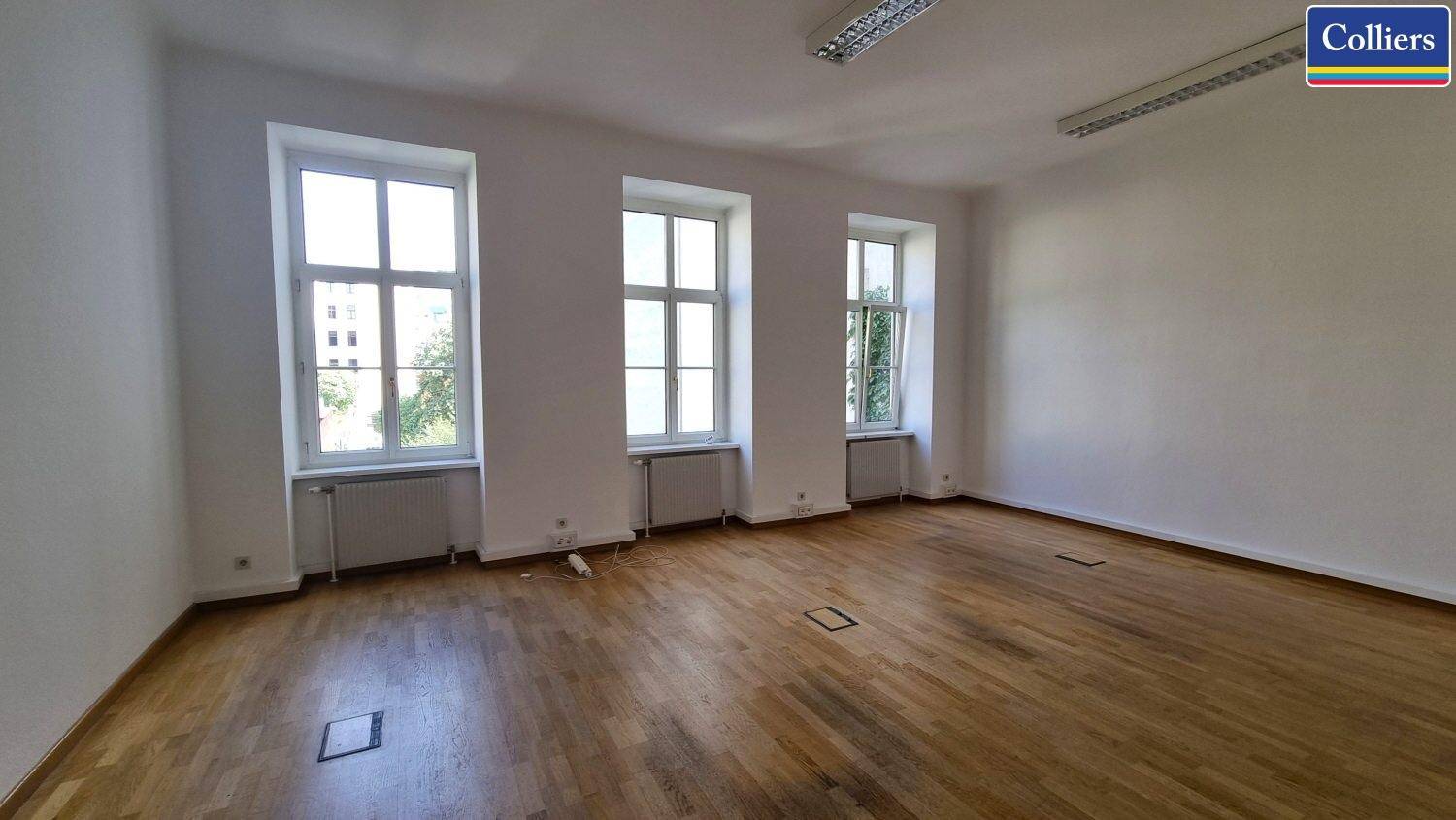 Ein heller Büroraum mit Holzboden, drei großen Fenstern und Bodentanks für Stromanschlüsse.