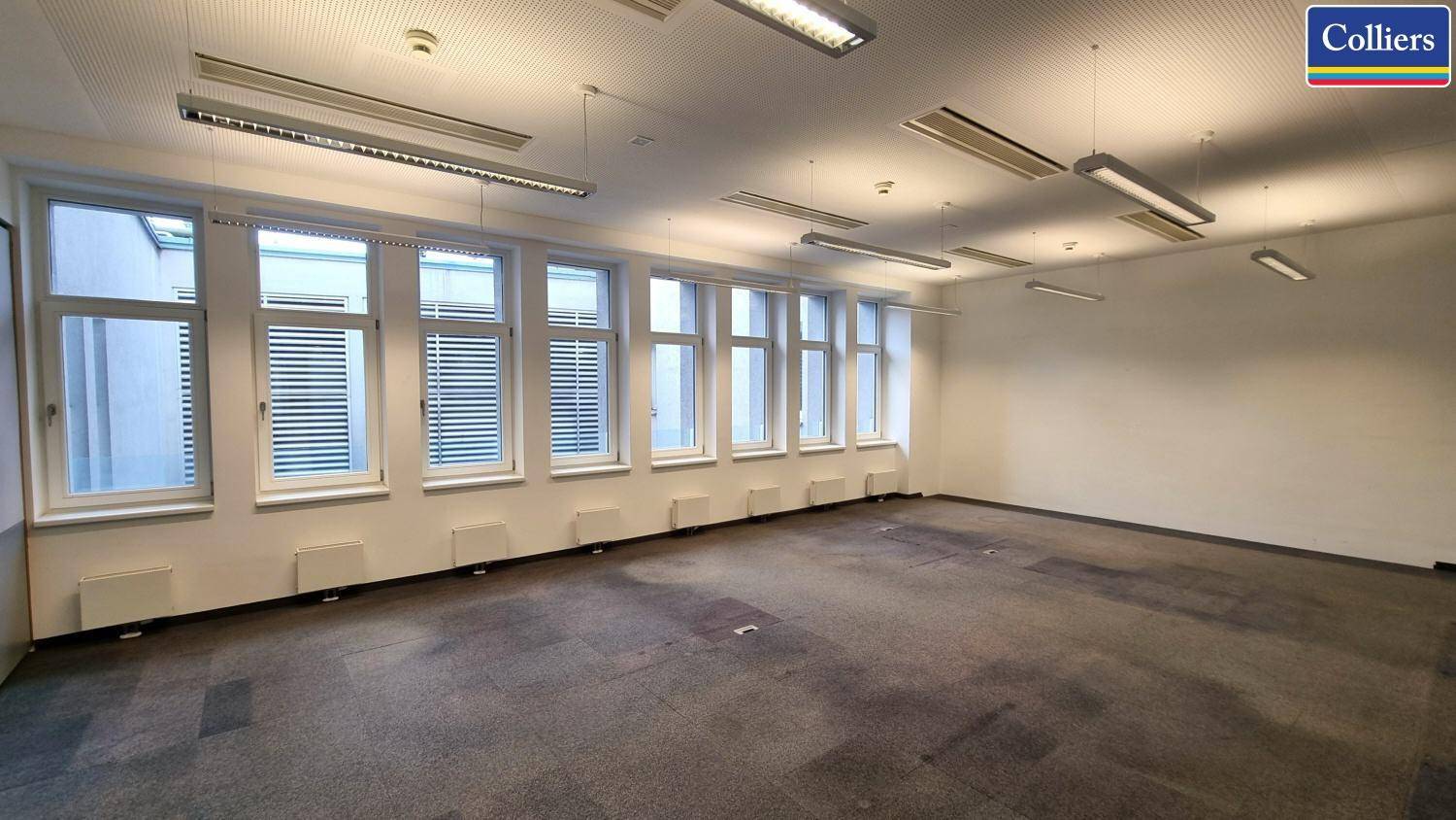 Geräumiges Büro mit einer Reihe von Fenstern, Teppichboden und Deckenbeleuchtung, ideal für Arbeitsbereiche.