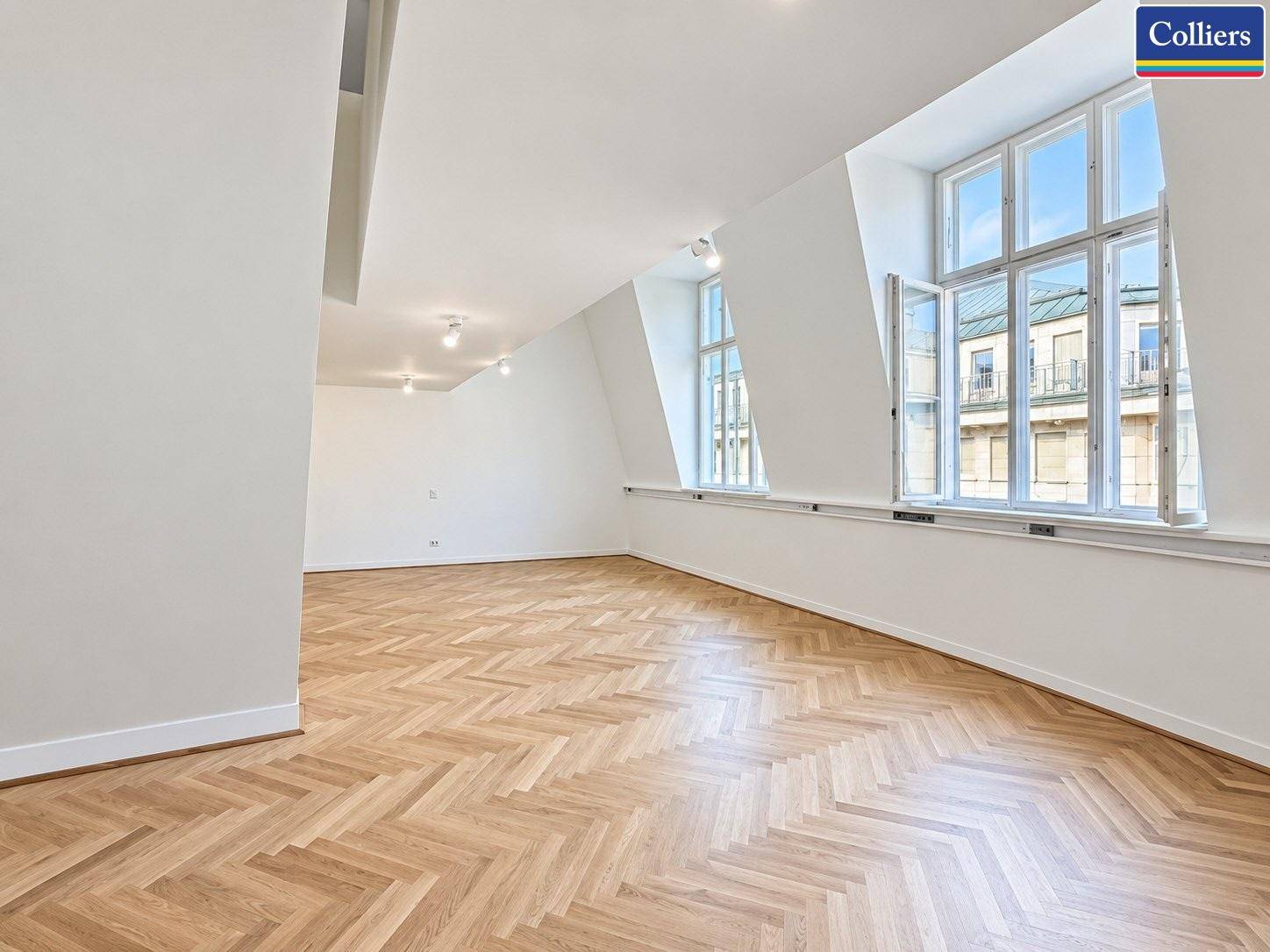 Geräumiger, heller Raum mit Fischgrätparkett, großen Fenstern und einer Dachschräge, die viel Tageslicht hereinlässt.