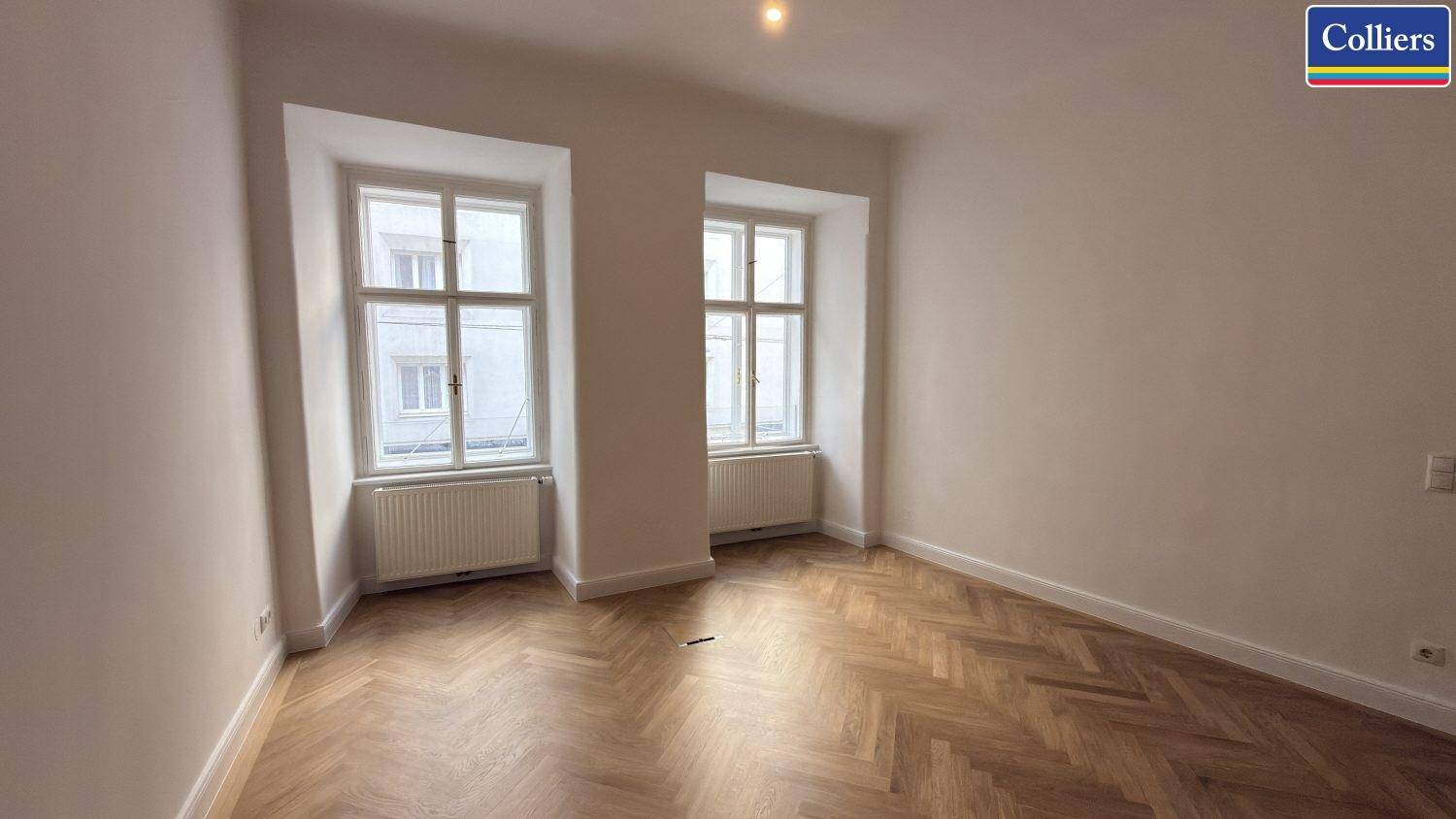 Heller Raum mit zwei Fenstern und Fischgrätparkett, bietet viel Tageslicht und eine angenehme Atmosphäre.