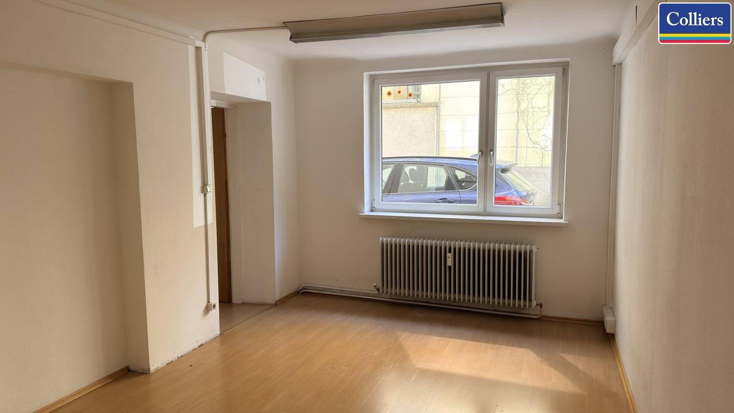 Heller Büroraum mit Laminatboden, einem Fenster zur Straße und einem Heizkörper für angenehme Wärme.
