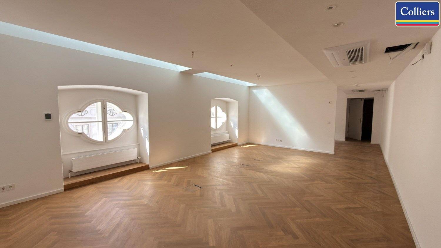 Heller Büroraum mit Fischgrätparkett, dekorativen Fenstern und Klimaanlage, die viel Tageslicht hereinlassen.