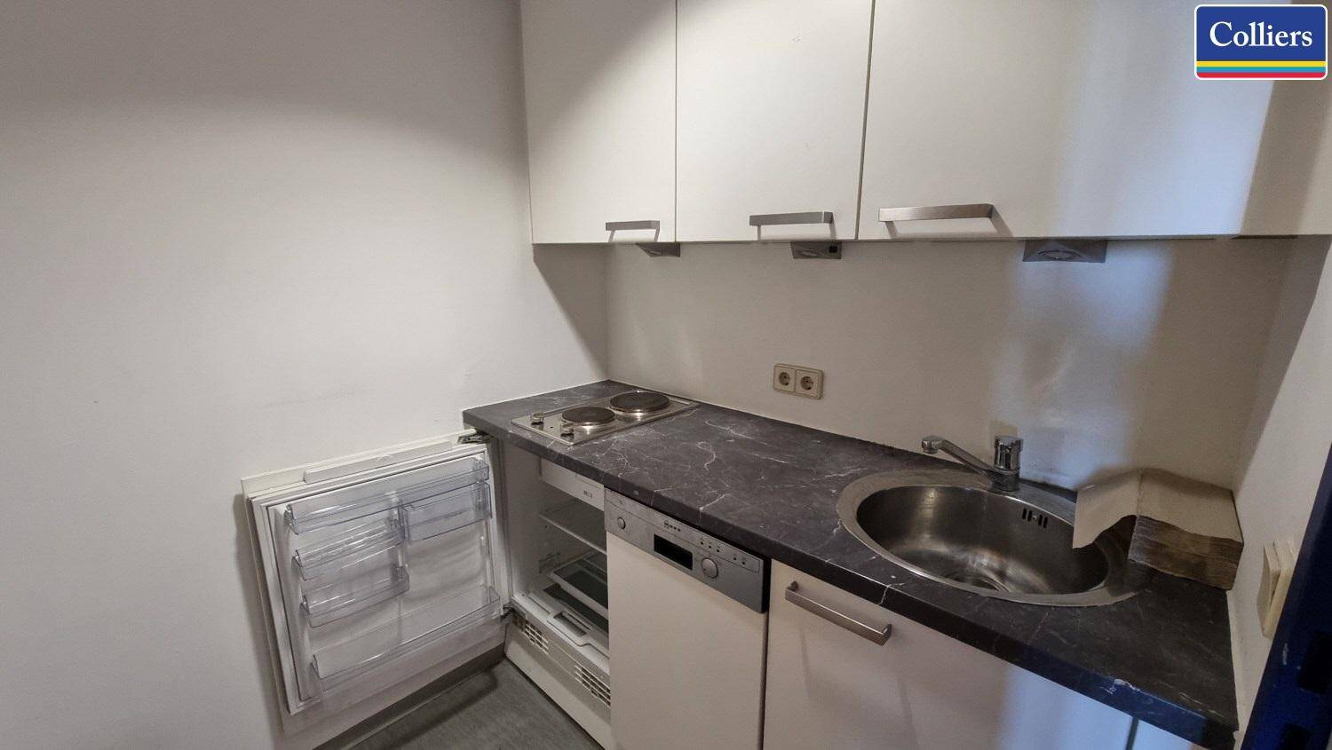 Kleine Küchenzeile mit Spüle, Kochplatten, Kühlschrank und weißen Hängeschränken.