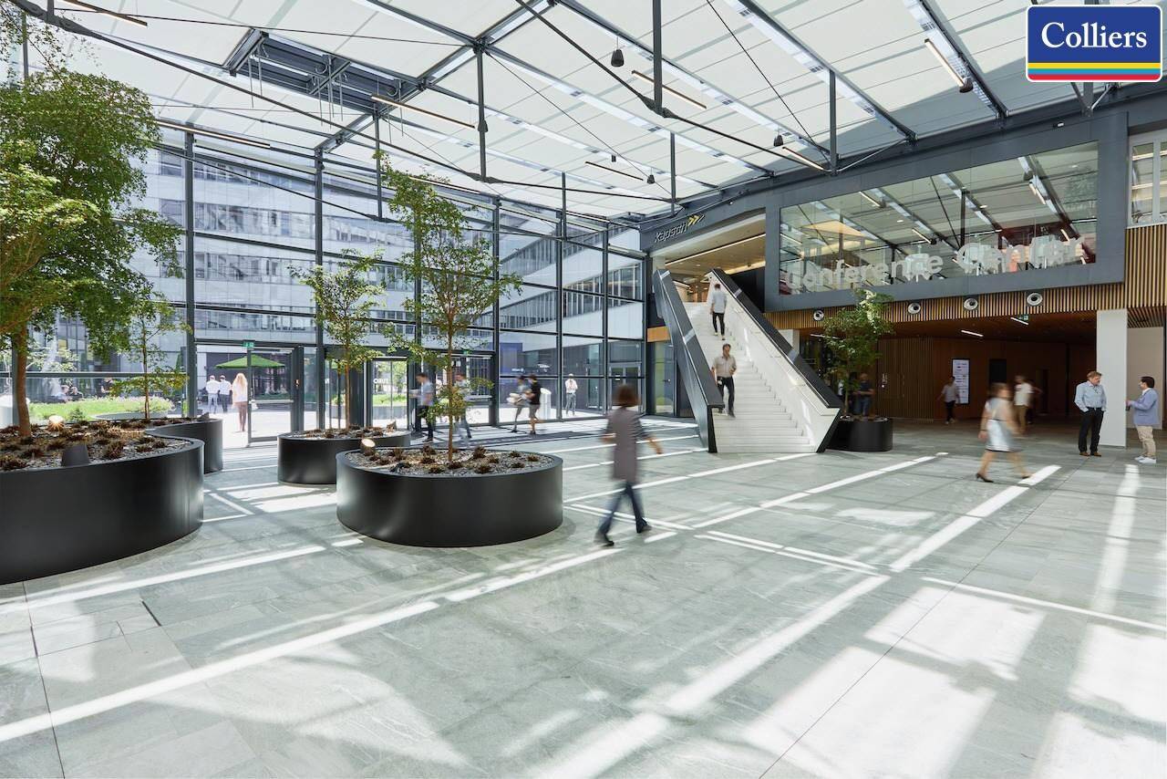 Großzügige, lichtdurchflutete Lobby mit hohen Glasfassaden, modernen Pflanzenkübeln und einer breiten Treppe.