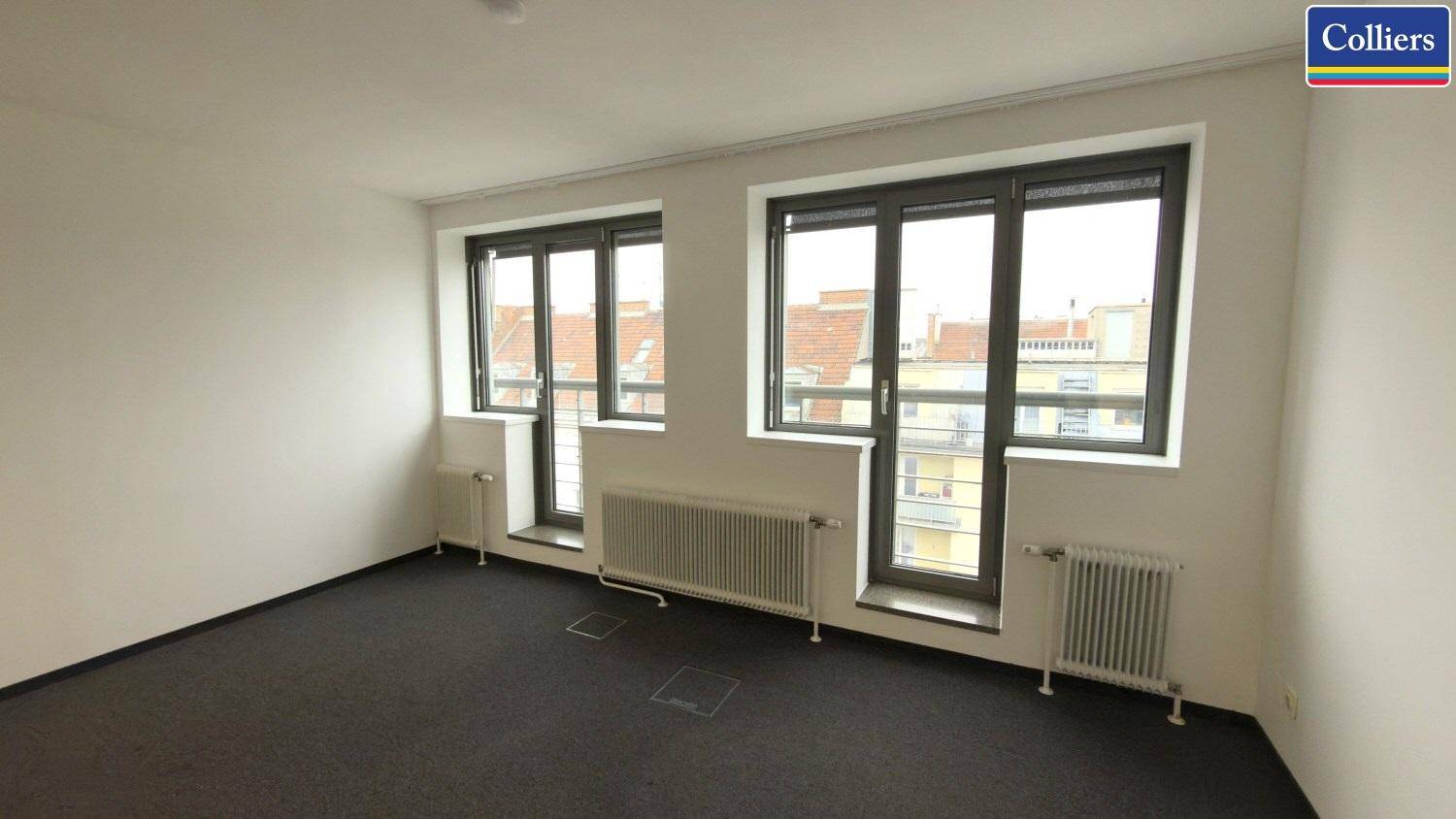 Geräumiges Büro mit dunklem Teppichboden, großen Fenstern und Heizkörpern, ideal für Arbeitsbereiche.