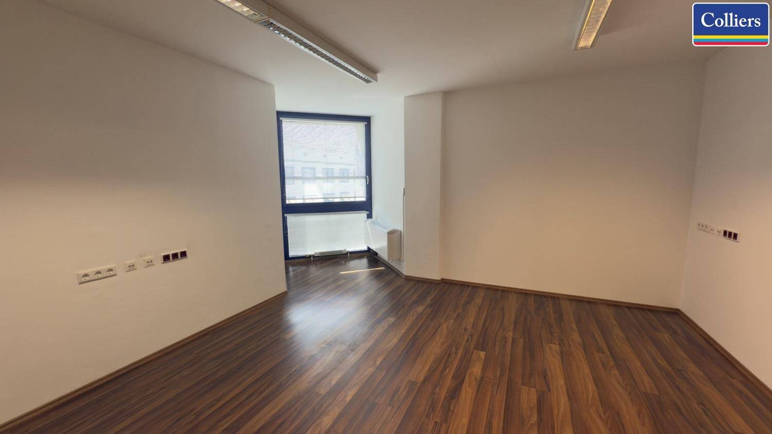 Bürofläche mit Holzboden und einem Fenster, das Tageslicht hereinlässt und einen Blick auf ein Gebäude bietet.