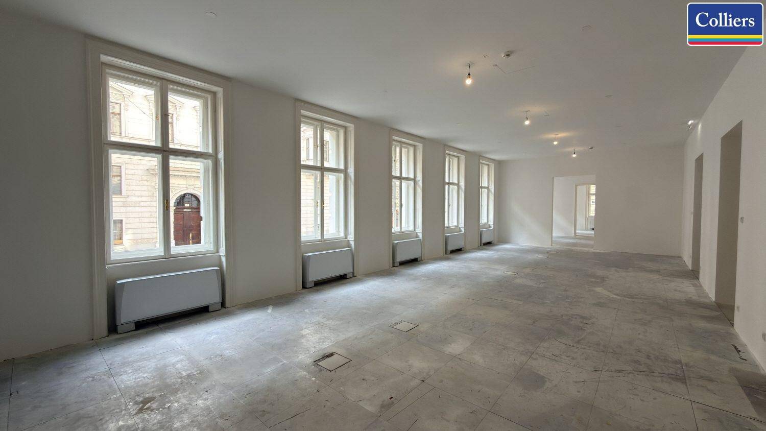 Geräumiger, unmöblierter Büroraum mit hohen Fenstern und hellem Steinboden, bereit für den Ausbau.