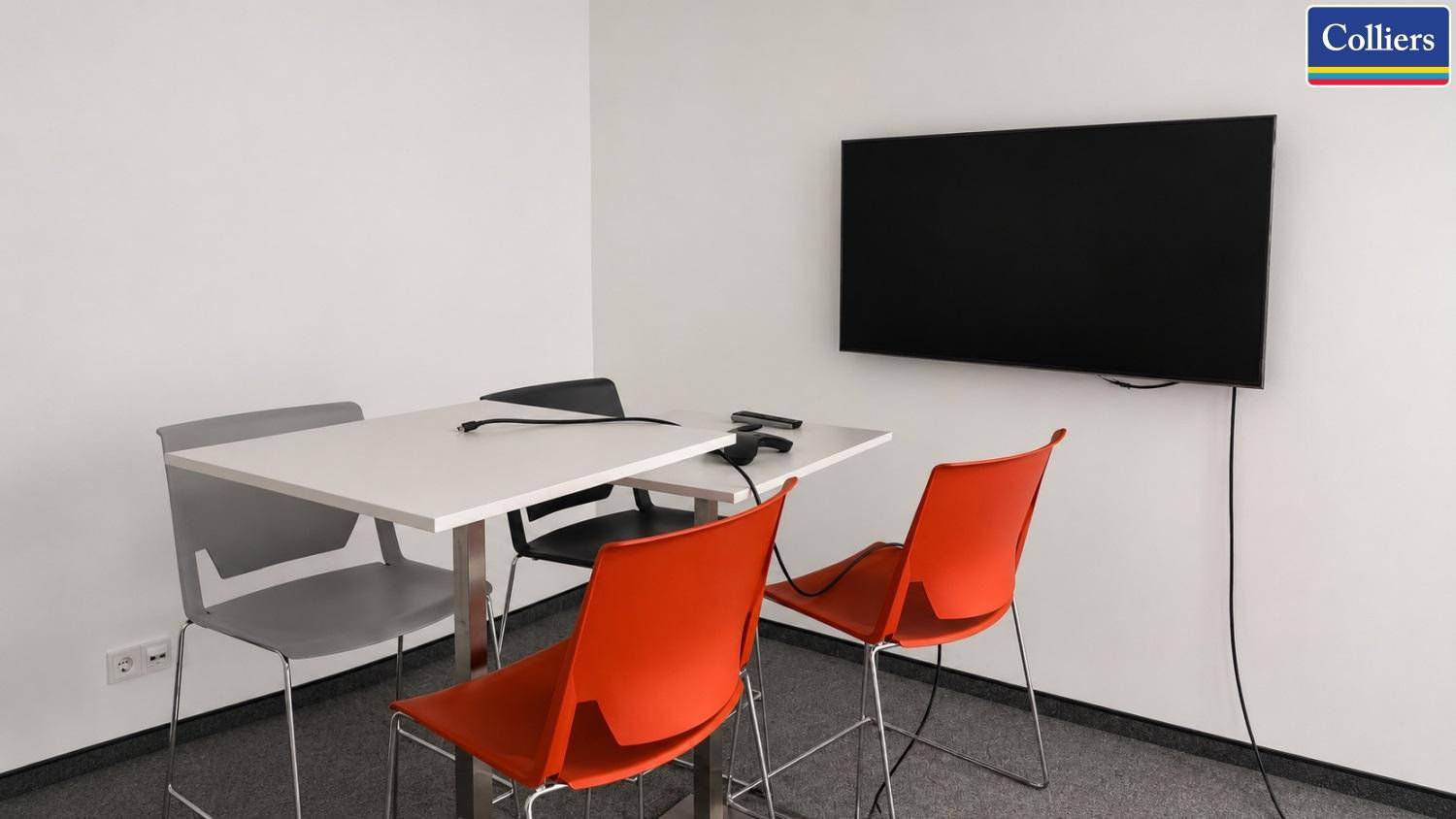 Kleiner Besprechungsraum mit Tisch, bunten Stühlen und Wandfernseher, ideal für kurze Team-Meetings.
