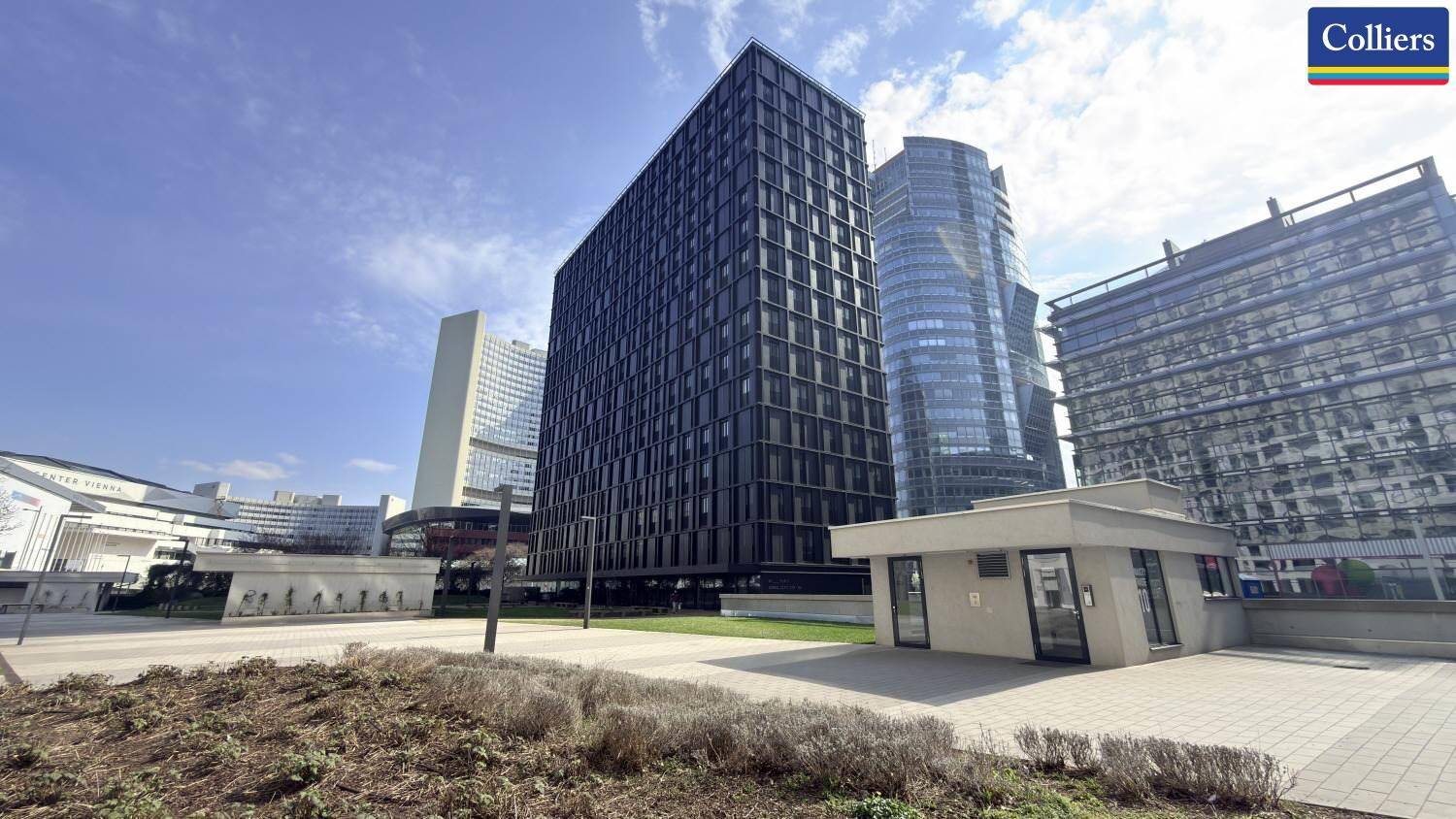 Panoramablick auf ein modernes Hochhaus mit dunkler Fassade inmitten einer städtischen Umgebung mit weiteren Gebäuden.