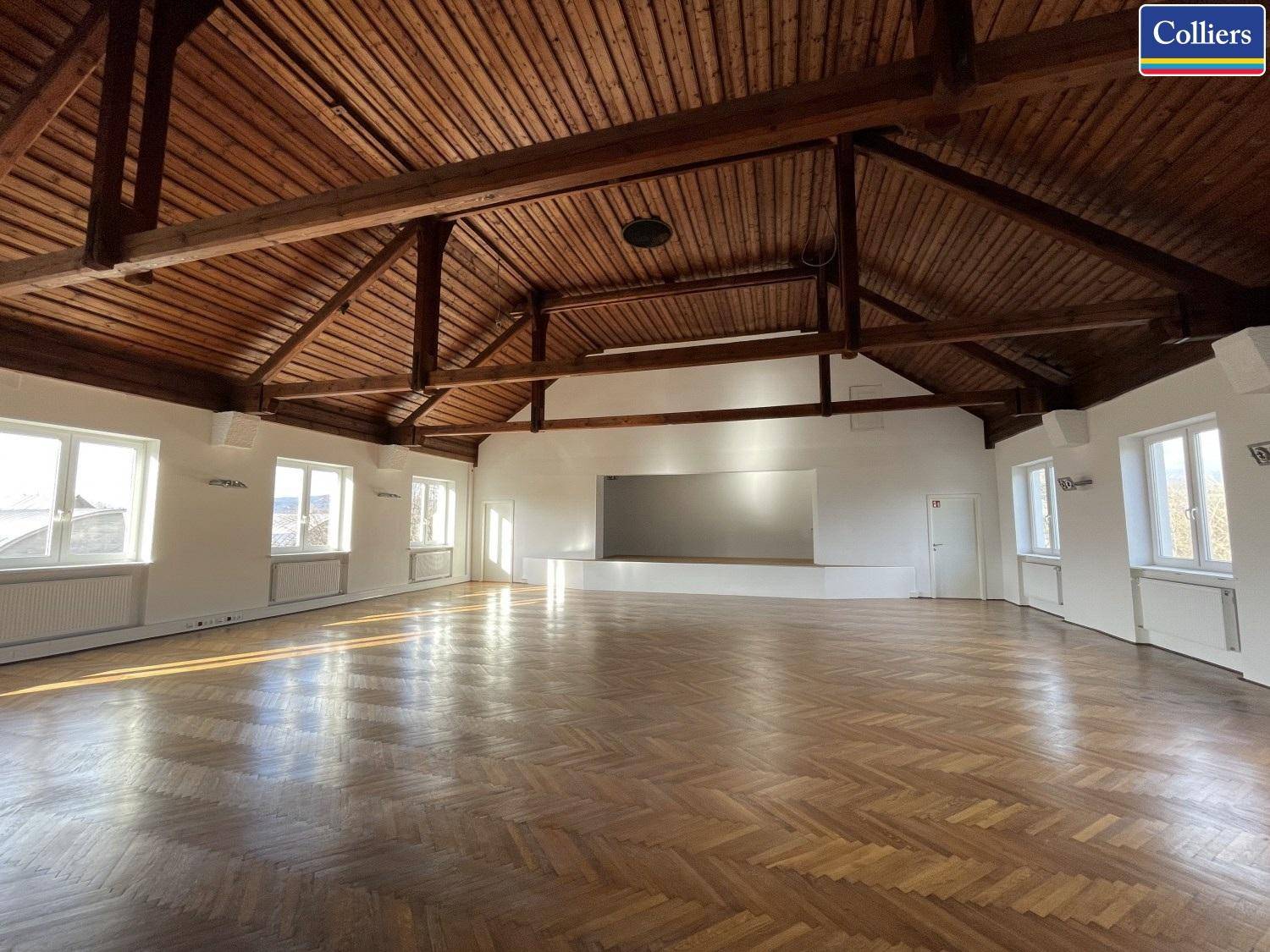 Großer Saal mit Holzbalkendecke, Fischgrätparkett und Bühne, ideal für Veranstaltungen.
