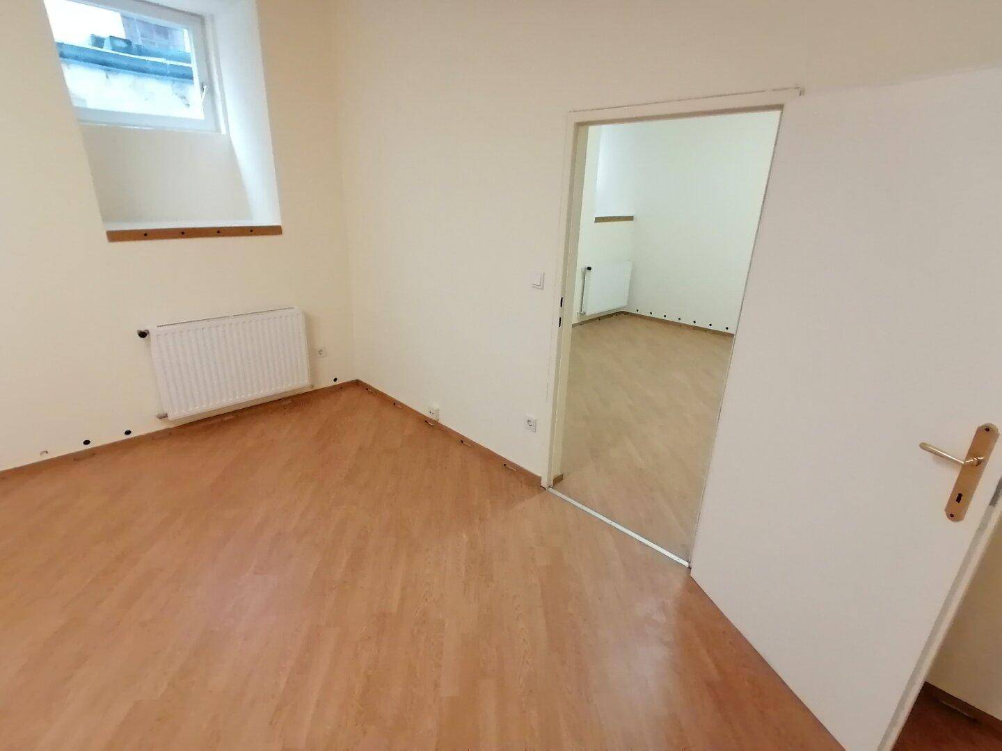 Kleinerer, heller Raum mit Laminatboden, einem Fenster und offener Tür, ideal als Schlafzimmer oder Büro.