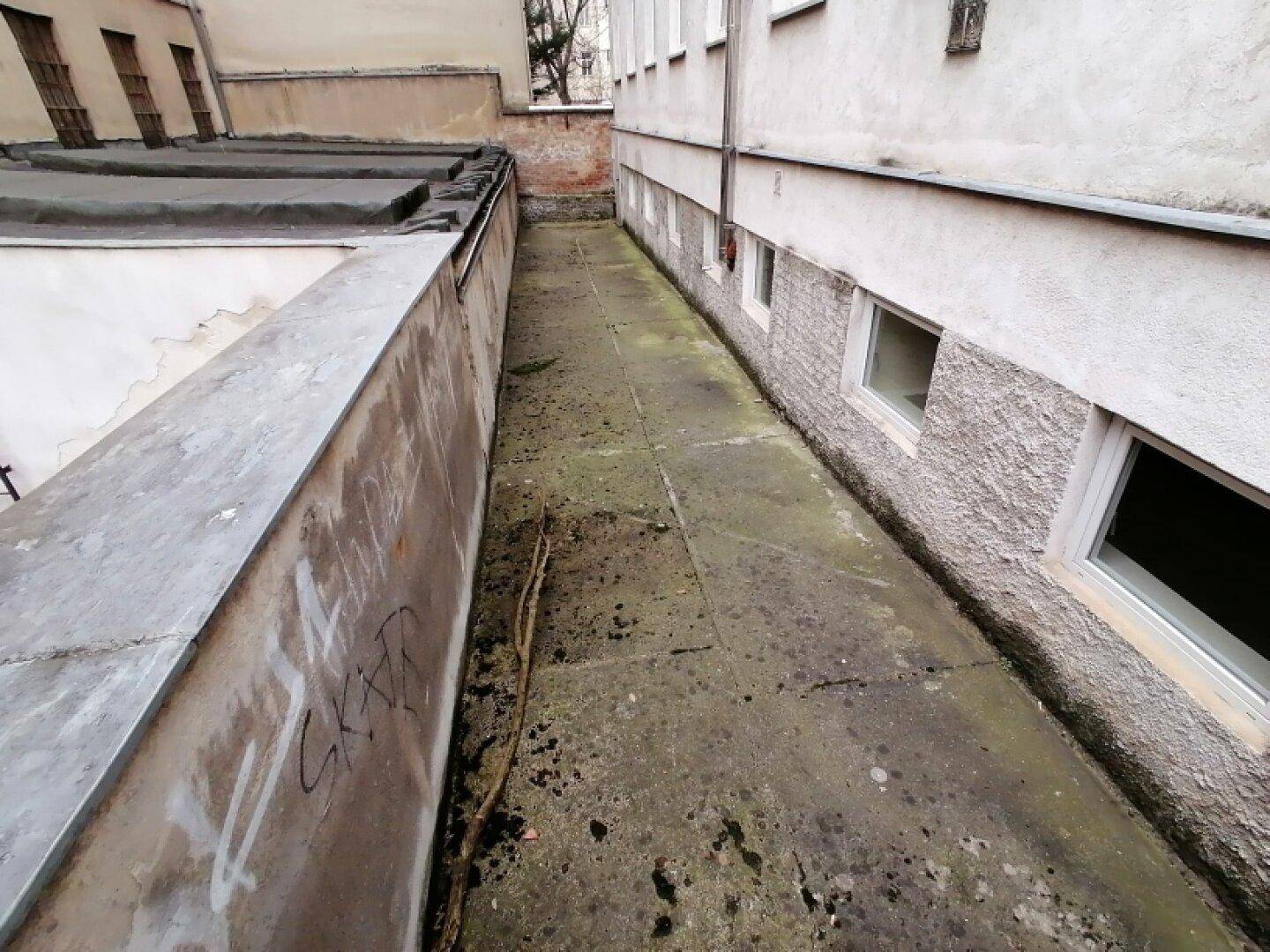 Ungepflegter Außenbereich zwischen Gebäuden mit schmalem Durchgang und sichtbaren Kellerfenstern, typisch für einen Altbau.