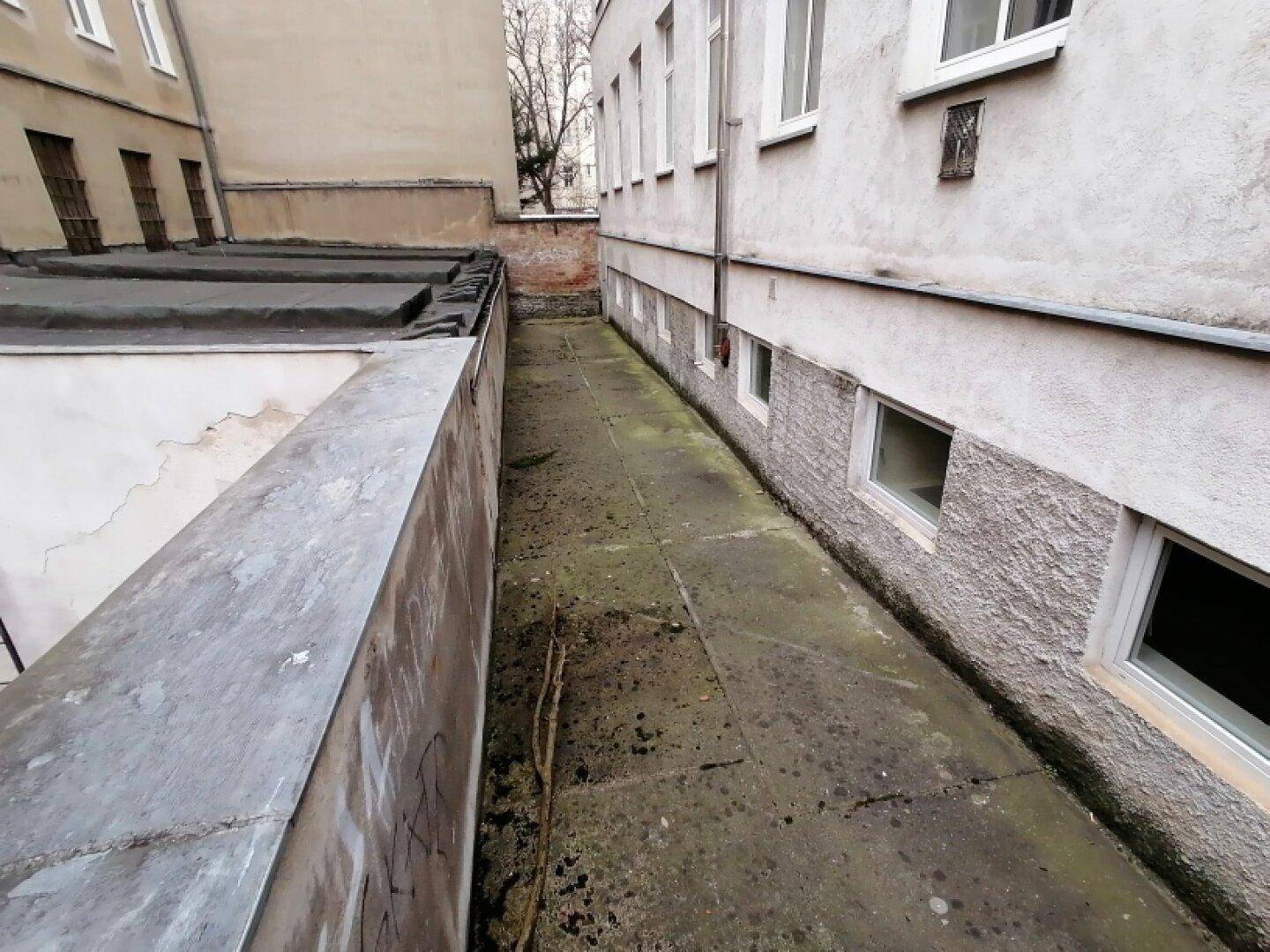 Schmaler, ungepflegter Außenbereich zwischen Gebäuden mit Betonboden und sichtbaren Kellerfenstern eines Altbaus.