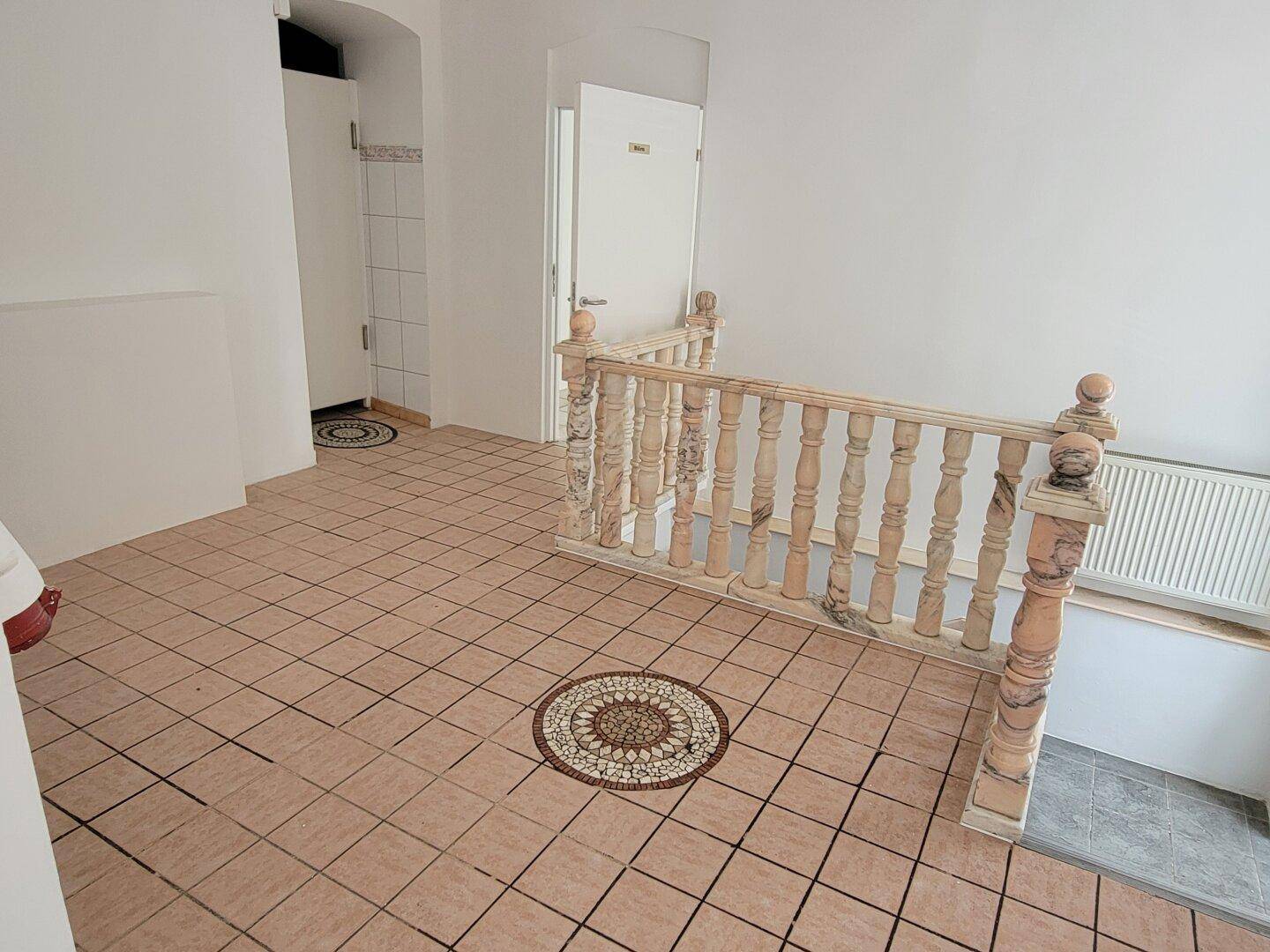 Flur mit gefliestem Boden, dekorativem Mosaik und Holzgeländer, das zu einem weiteren Bereich führt.