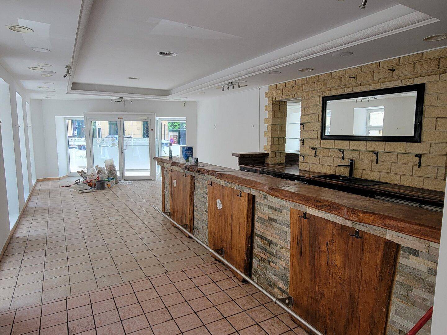 Geräumiger Barbereich mit langer Theke aus Holz und Stein, gefliestem Boden und großen Fenstern zur Straße.