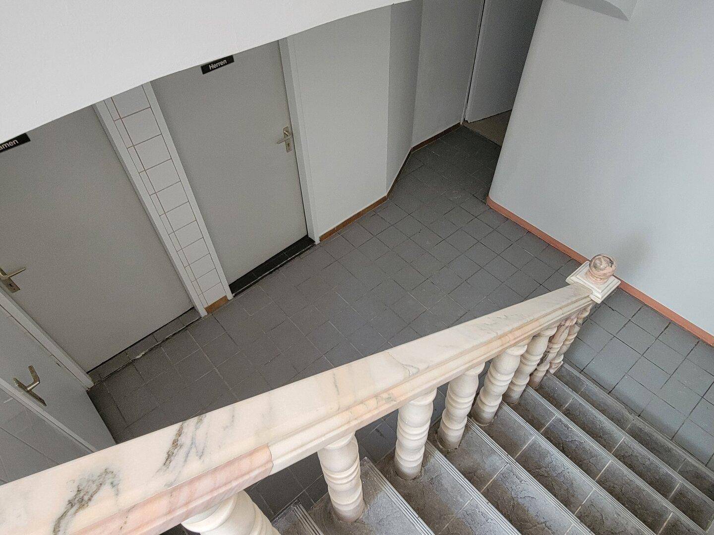 Treppenhaus mit Marmorgeländer und gefliestem Boden, das zu den Toiletten im unteren Bereich führt.