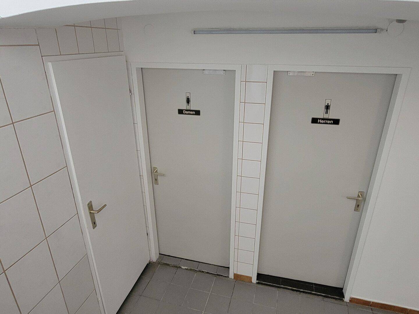 Toilettenbereich mit zwei Türen für Damen und Herren, weißen Fliesenwänden und grauem Fliesenboden.