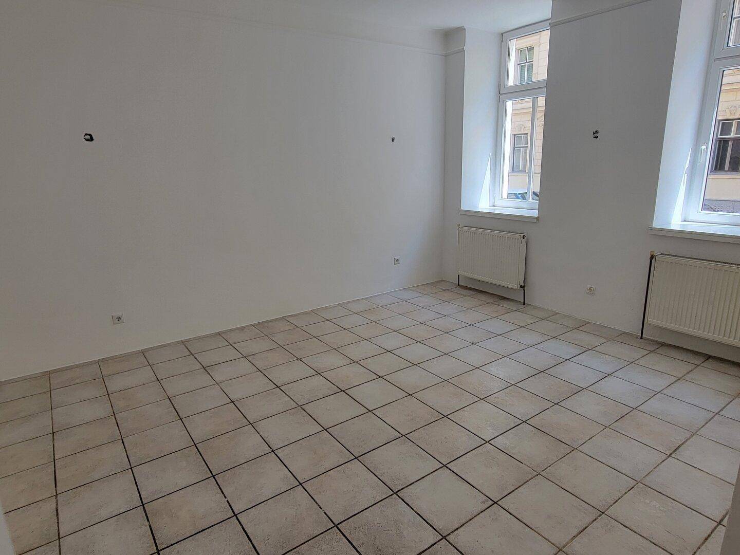 Leerer Raum mit weißen Wänden, gefliestem Boden und zwei Fenstern mit Heizkörpern, die viel Licht spenden.