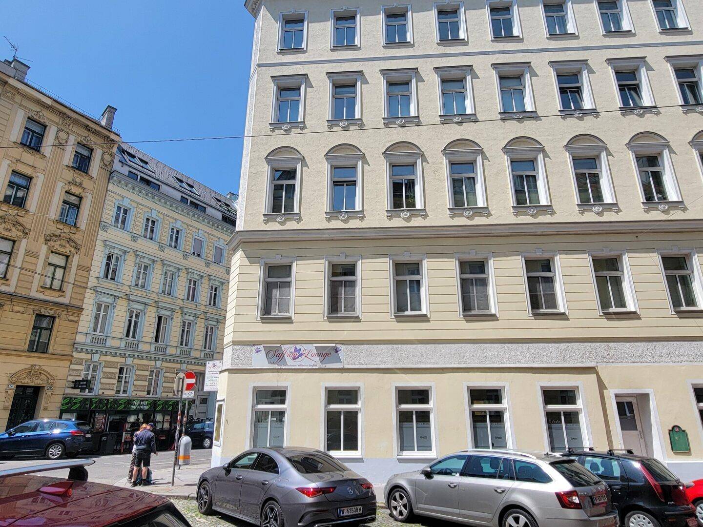 Ansicht des Eckgebäudes von der Straße aus mit geparkten Autos und umliegenden Gebäuden.