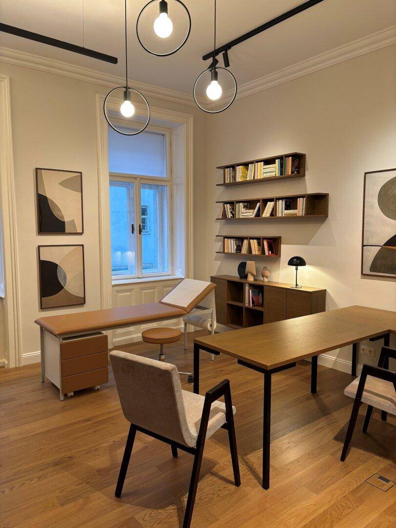 Helles Behandlungszimmer mit Schreibtisch, Untersuchungsliege, Bücherregalen und großen Fenstern.