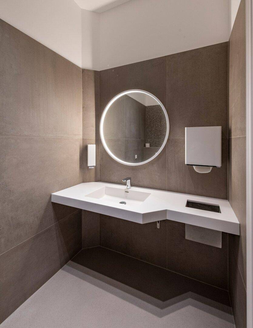 Modernes Badezimmer mit großem Waschbecken, beleuchtetem Rundspiegel und grauen Fliesen.