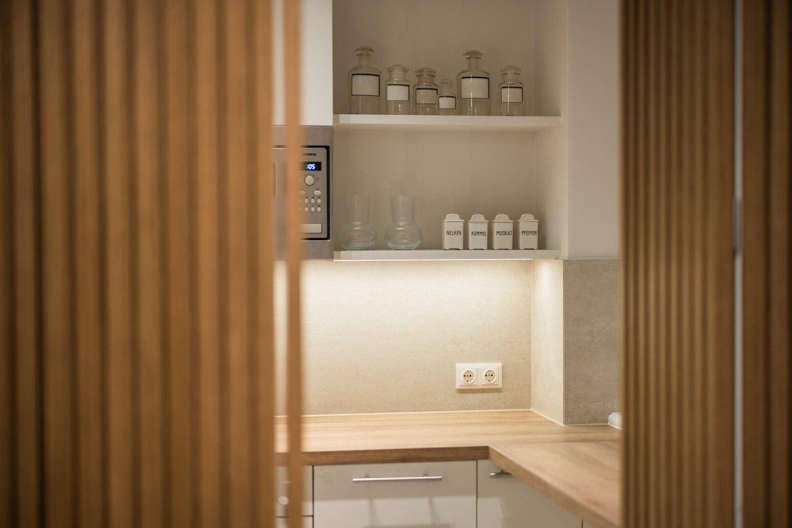 Kleine Teeküche mit Mikrowelle, offenen Regalen und Holztheke, geschickt hinter Lamellen versteckt.