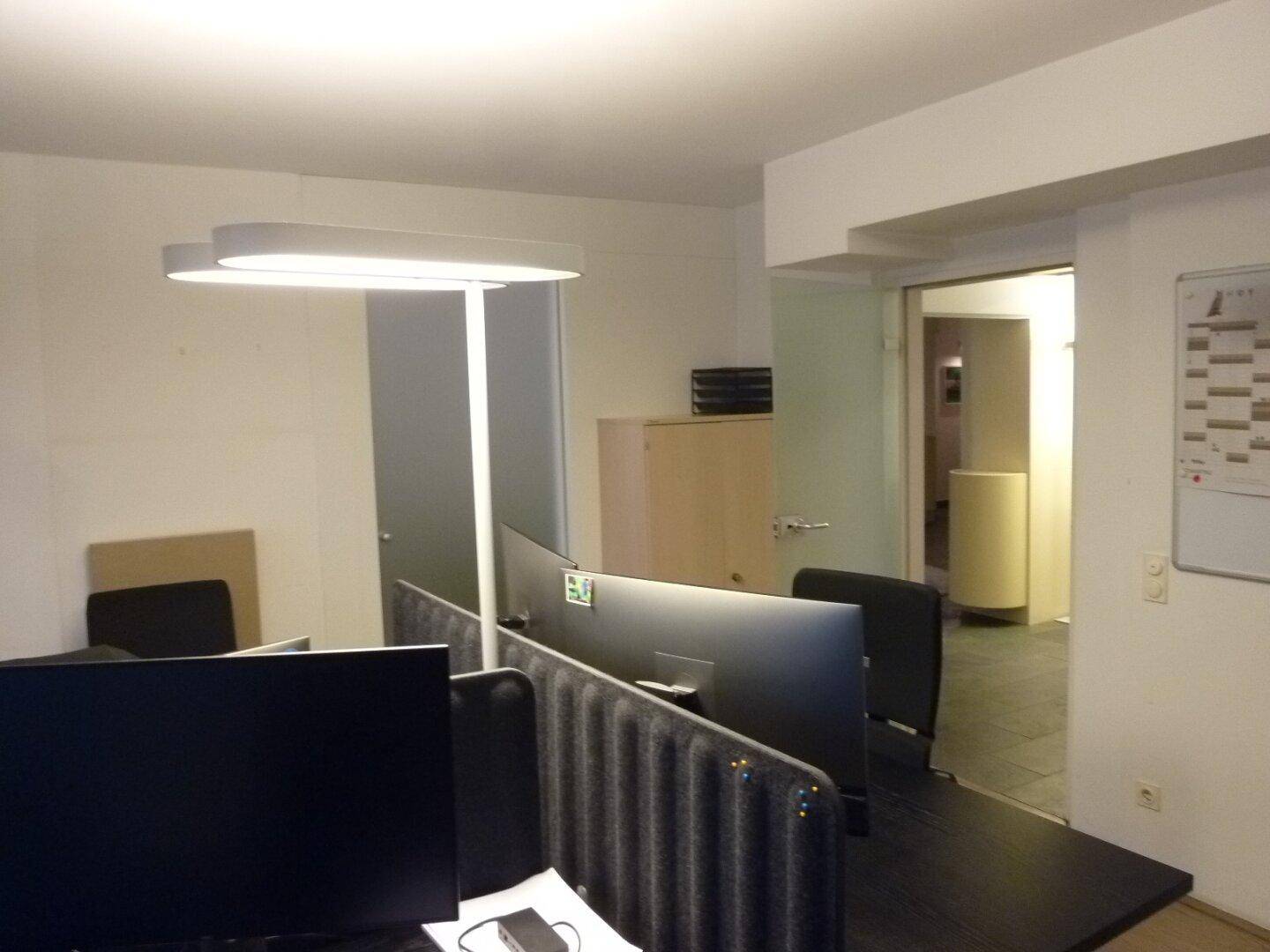 Moderner Büroarbeitsplatz mit Trennwänden, ergonomischen Stühlen und Blick in den Flur.