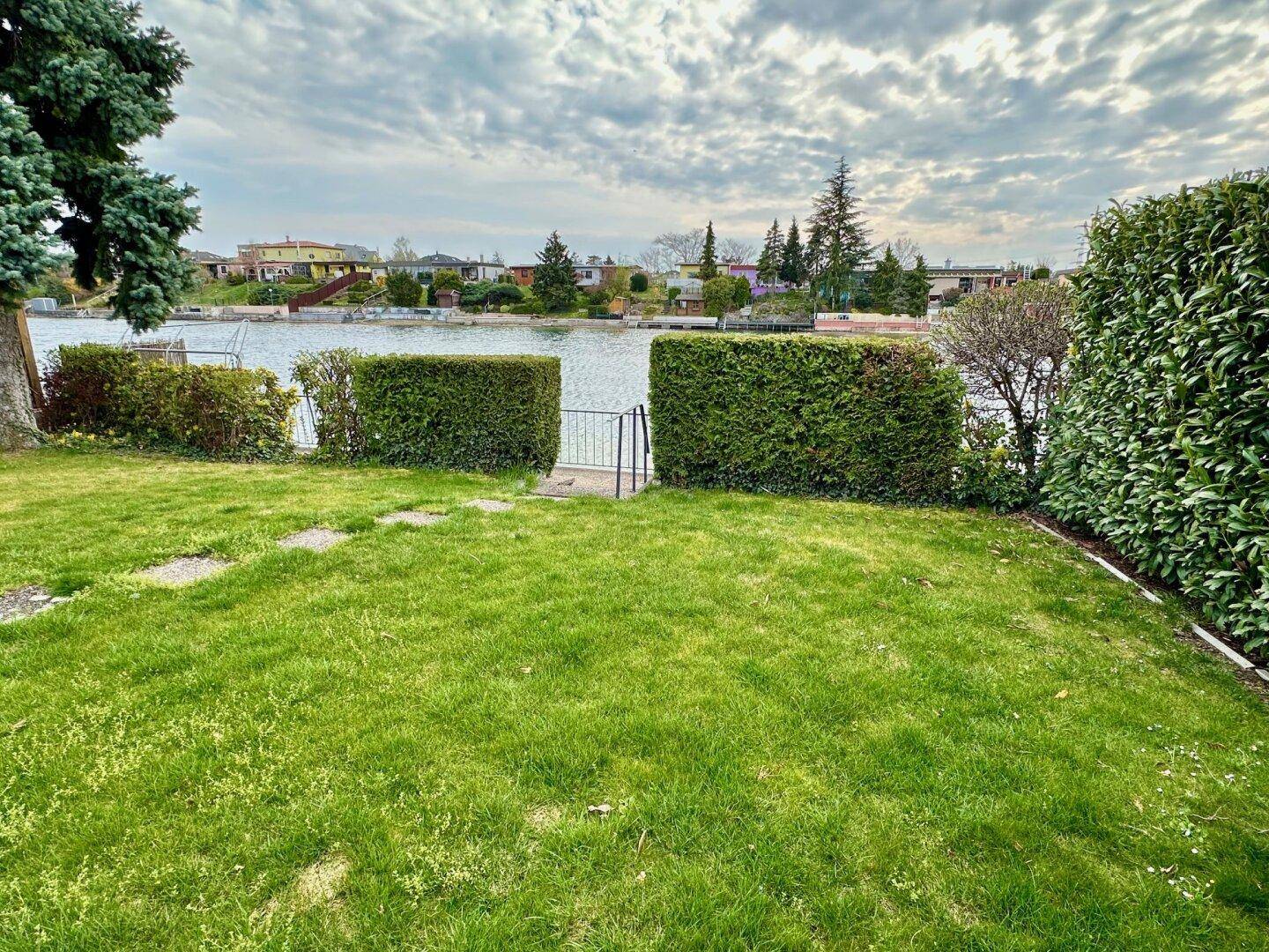 Weitläufiger Garten mit Rasenfläche, Hecken und direktem Blick auf den See.