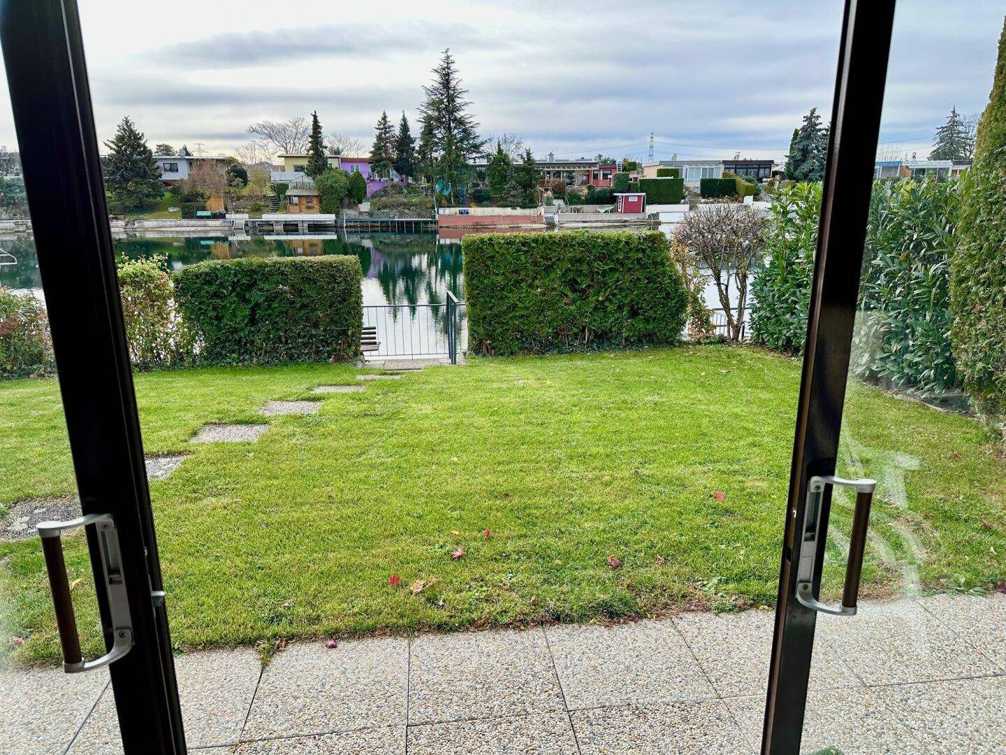 Blick aus dem Innenraum durch große Fenster auf den grünen Garten und den dahinterliegenden See.