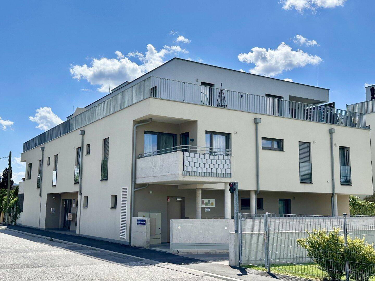 Modernes Mehrfamilienhaus mit heller Fassade, Balkonen und Dachterrasse unter blauem Himmel.