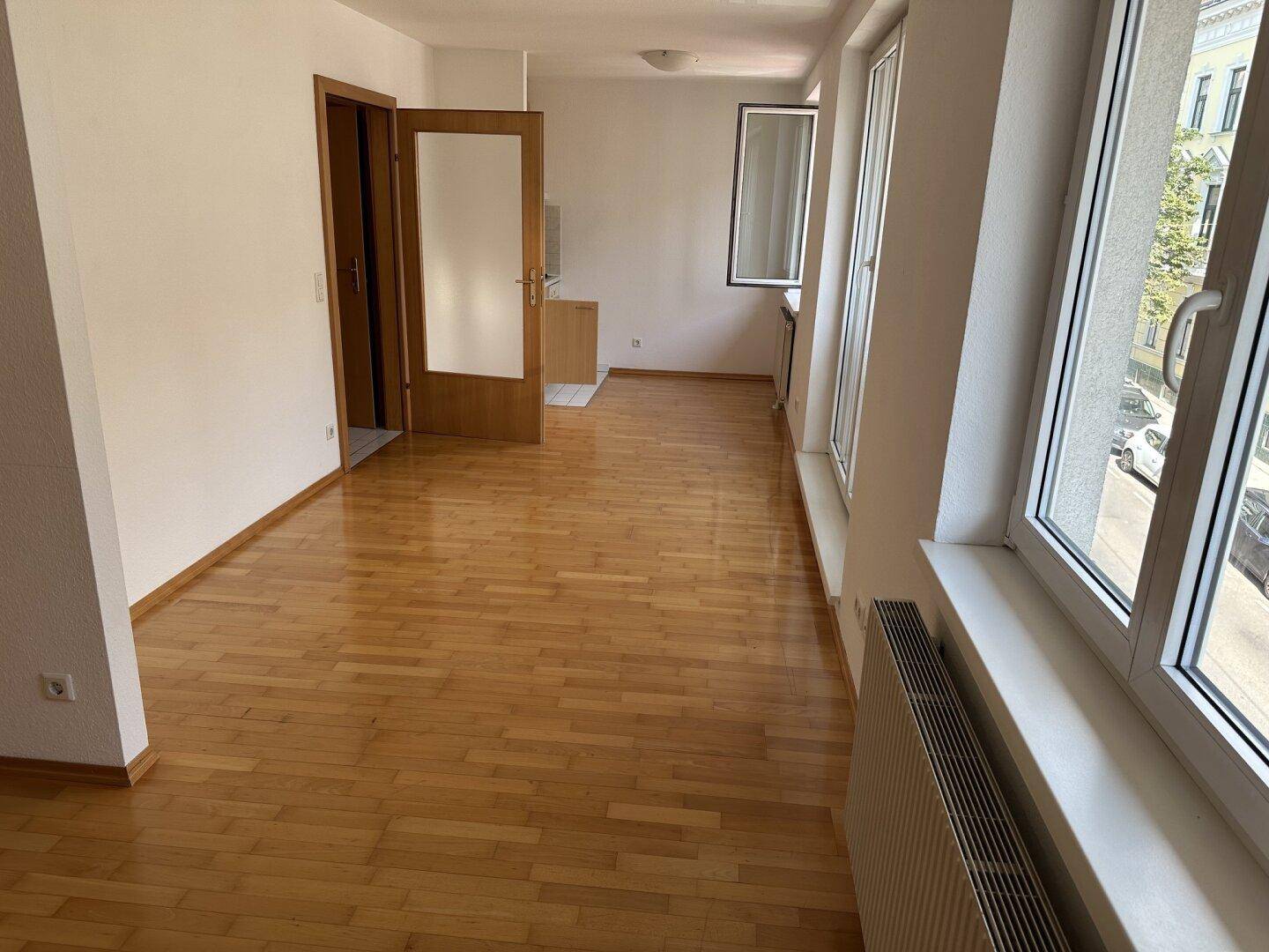 Heller Wohnraum mit Parkettboden, großen Fenstern und einer Tür zu einem weiteren Zimmer.
