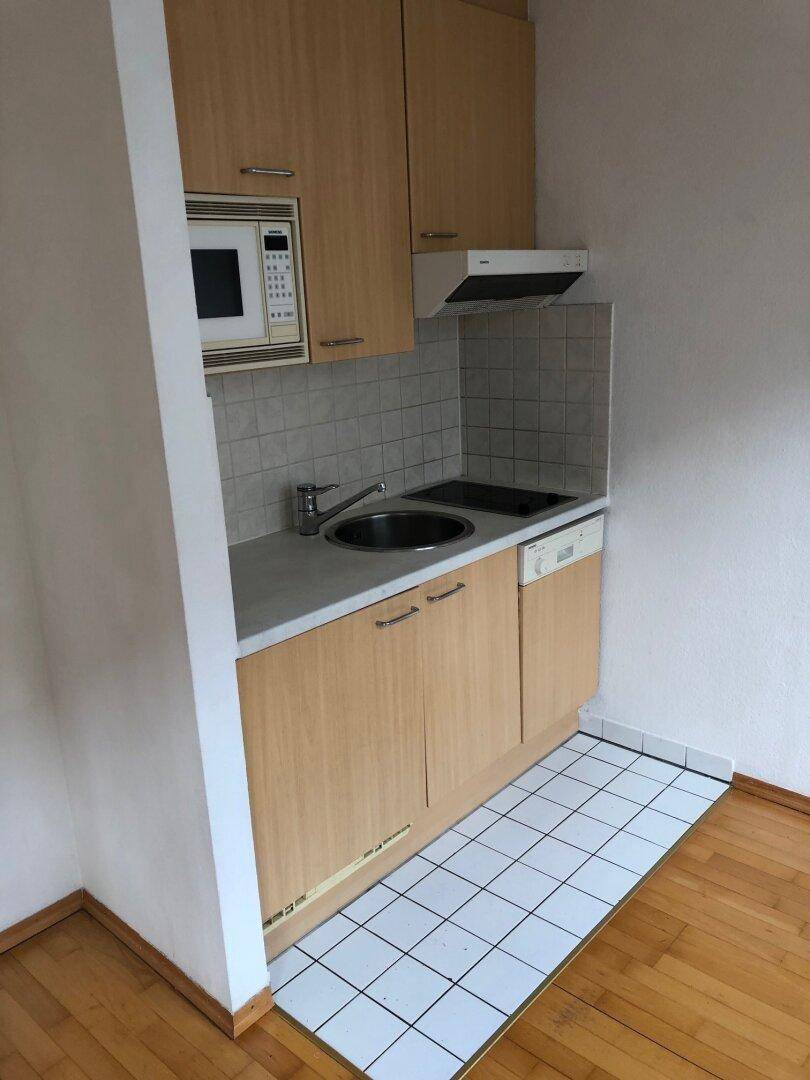 Kompakte Kochnische mit Holzschränken, Spüle, Kochfeld und Mikrowelle, ideal für kleine Wohnungen.