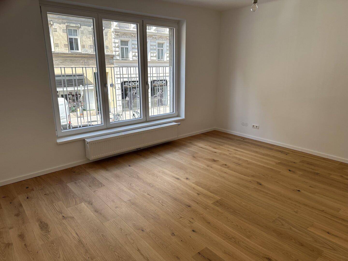 Geräumiges Zimmer mit Holzboden und großen Fenstern, die viel Tageslicht hereinlassen und einen Blick auf die Straße bieten.