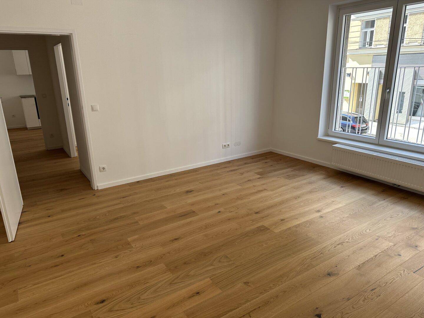 Heller Wohnbereich mit Holzboden und Zugang zu einem weiteren Raum, durch große Fenster gut belichtet.