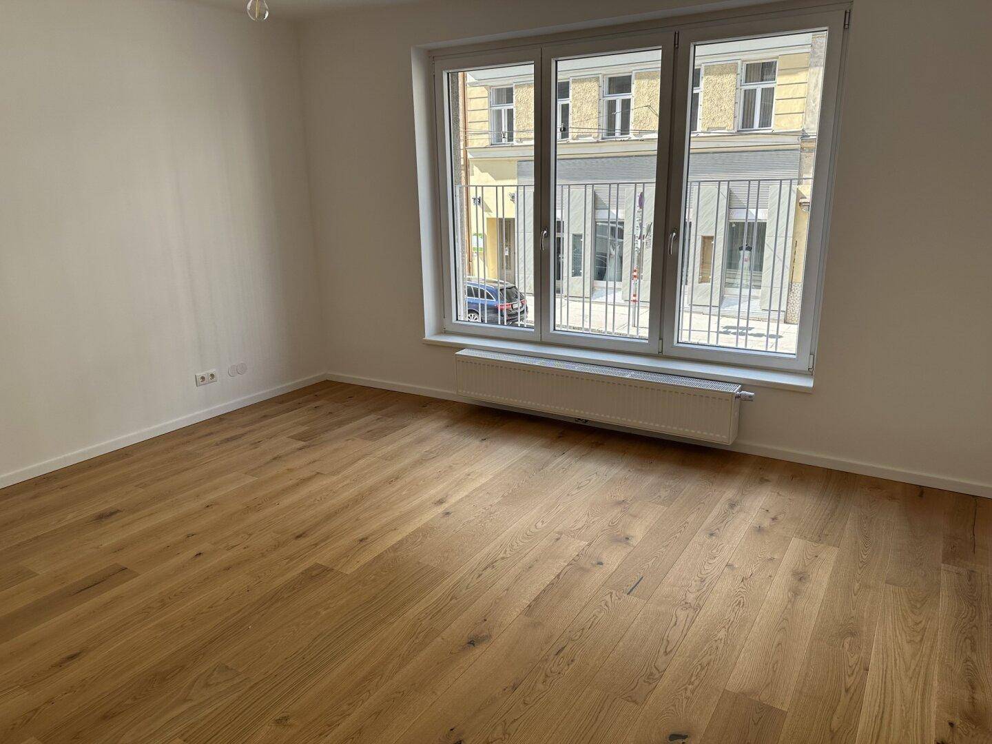 Lichtdurchflutetes Zimmer mit Holzboden und großen Fenstern, die einen Blick auf die gegenüberliegende Bebauung ermöglichen.