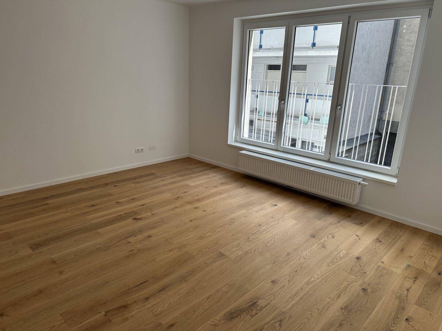 Helles Zimmer mit Holzboden und großen Fenstern, die einen Blick auf einen Innenhof oder eine Rückseite bieten.