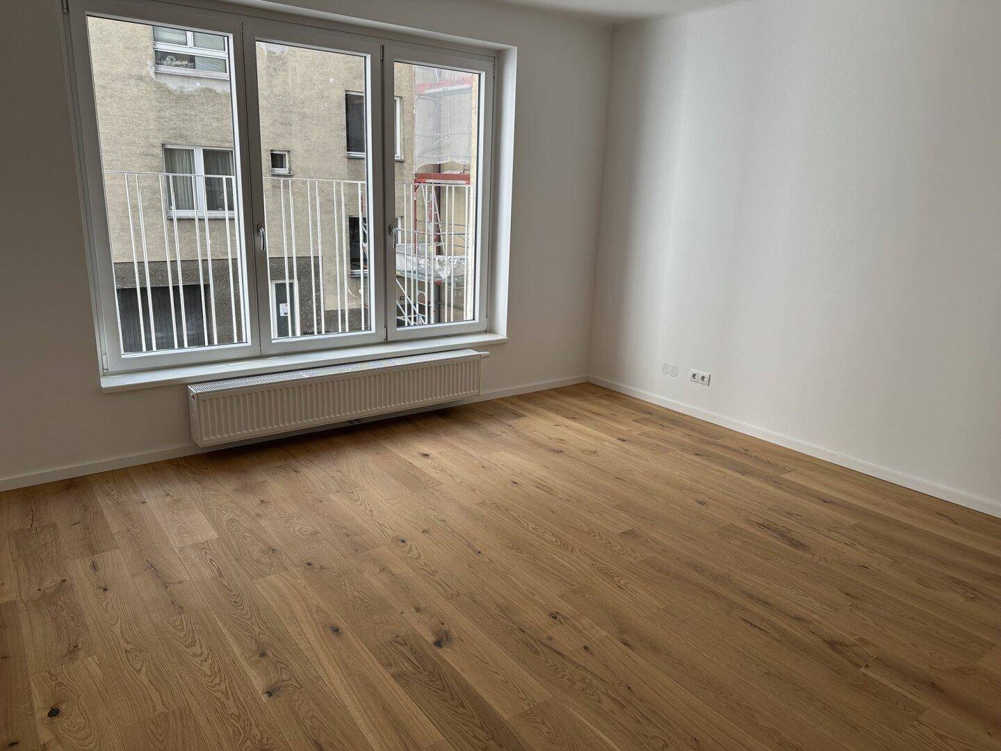 Geräumiges Zimmer mit Holzboden und großen Fenstern, die viel Tageslicht hereinlassen und einen Blick auf den Innenhof bieten.