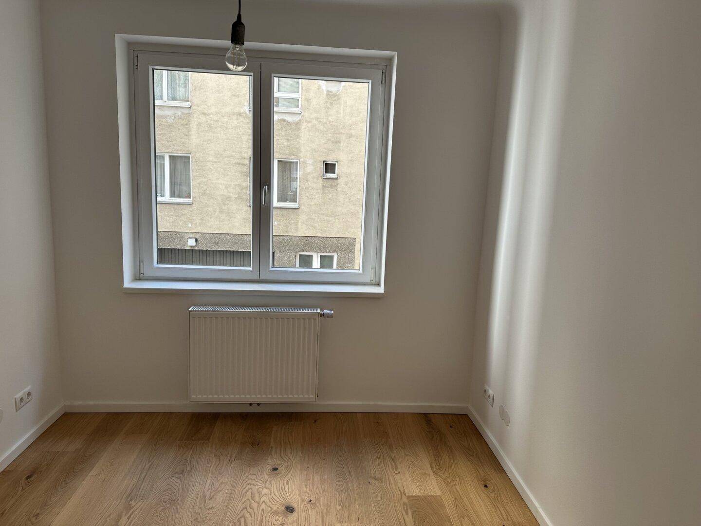 Helles Zimmer mit Holzboden und großem Fenster, das einen Blick auf den Innenhof bietet.